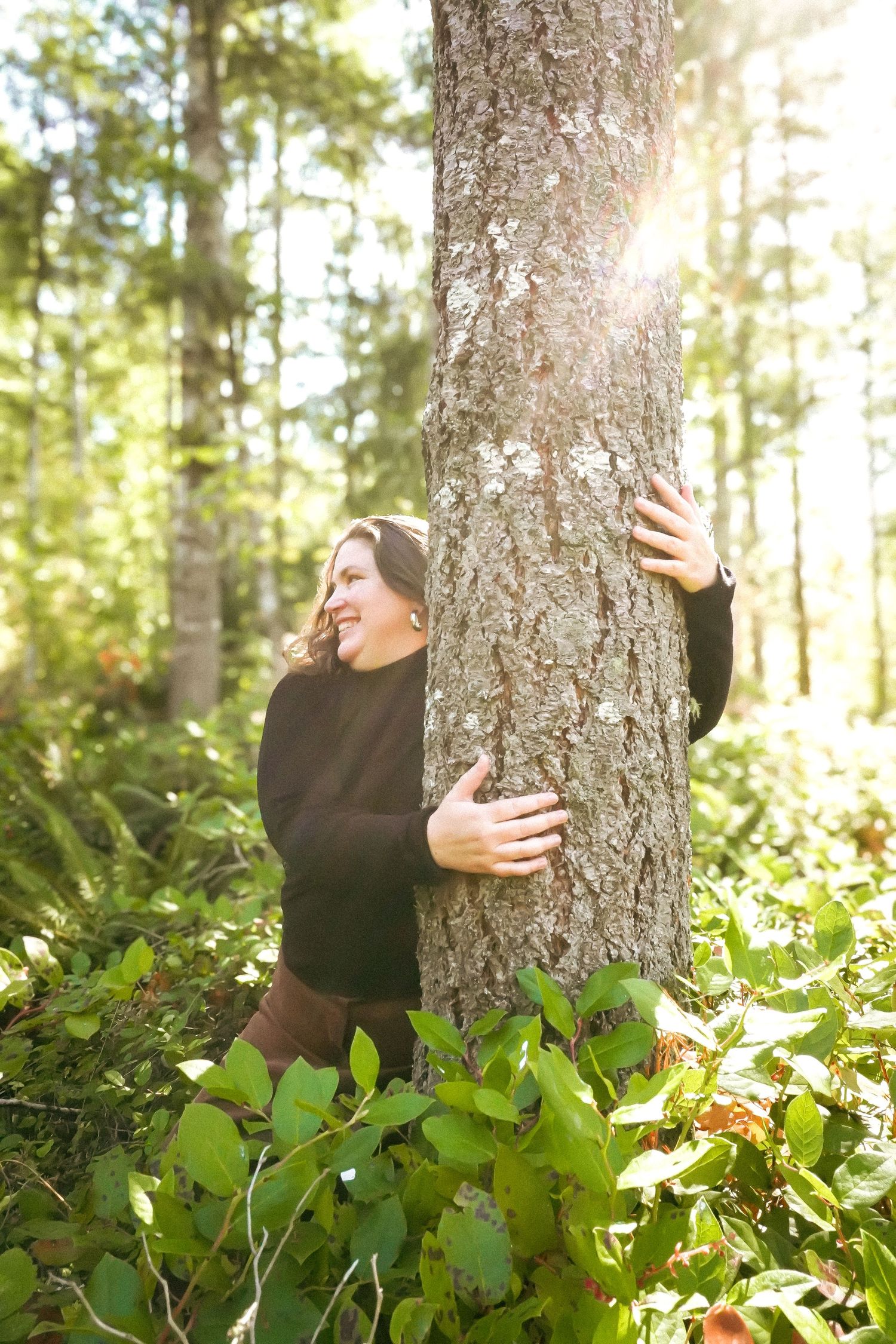 A smiling woman hugging a large tree trunk in a sunlit forest surrounded by green foliage.