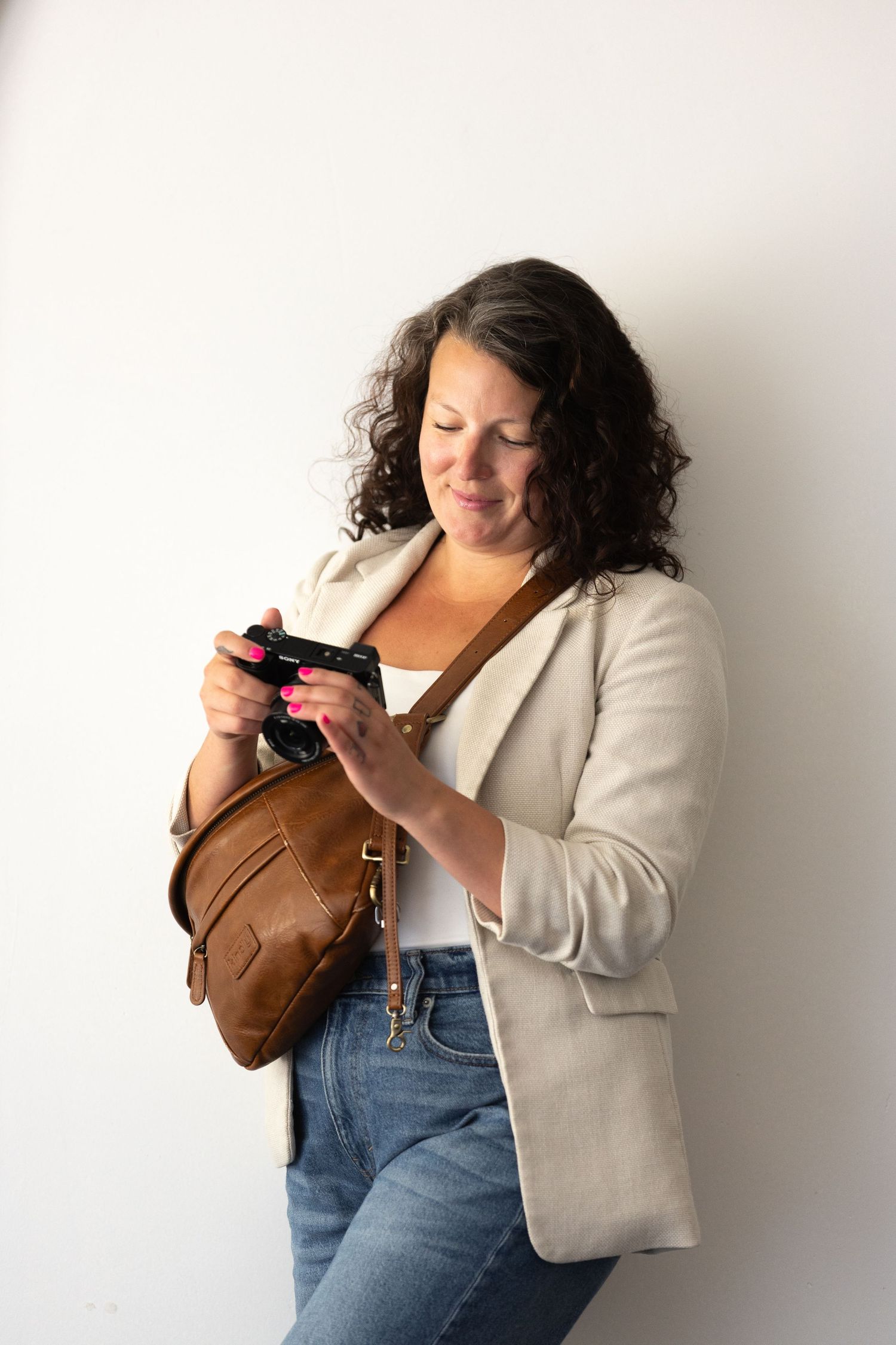 Woman in beige blazer reviewing photos on Sony camera with brown leather crossbody bag against white studio wall.