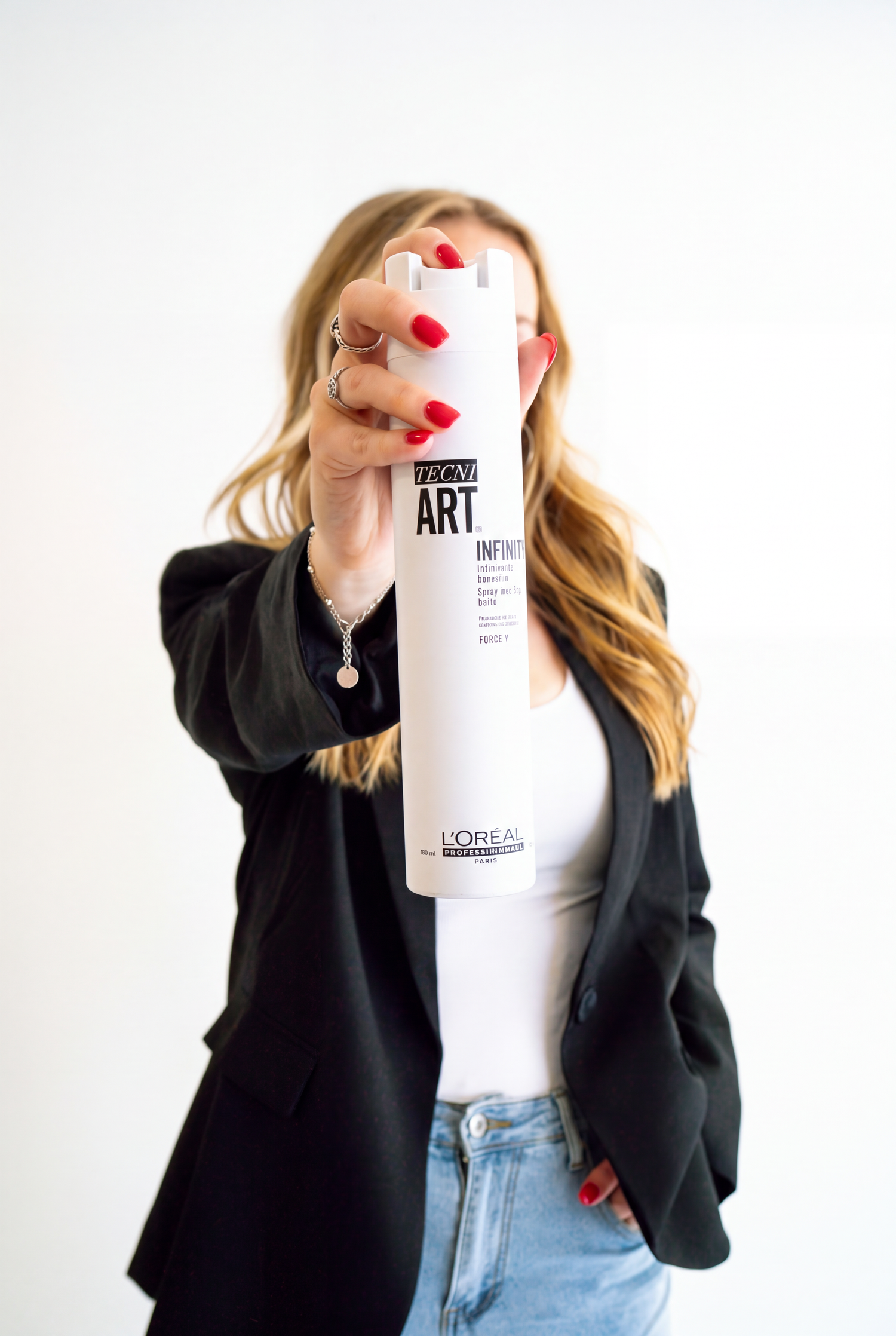 Woman in a black blazer and jeans holds a white L'Oréal Professionnel Tecni.Art hairspray can toward the camera, red nails visible.