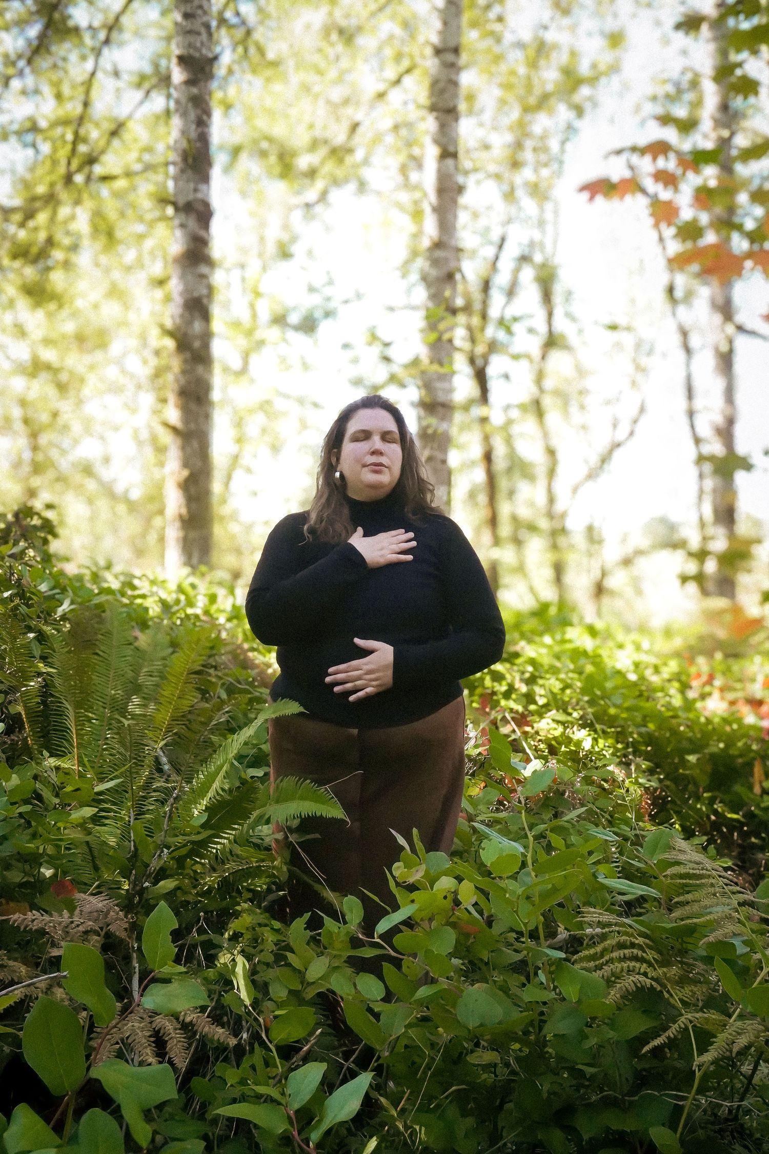 A woman in a black turtleneck stands among lush green ferns in a sunlit forest, hands on chest and belly.