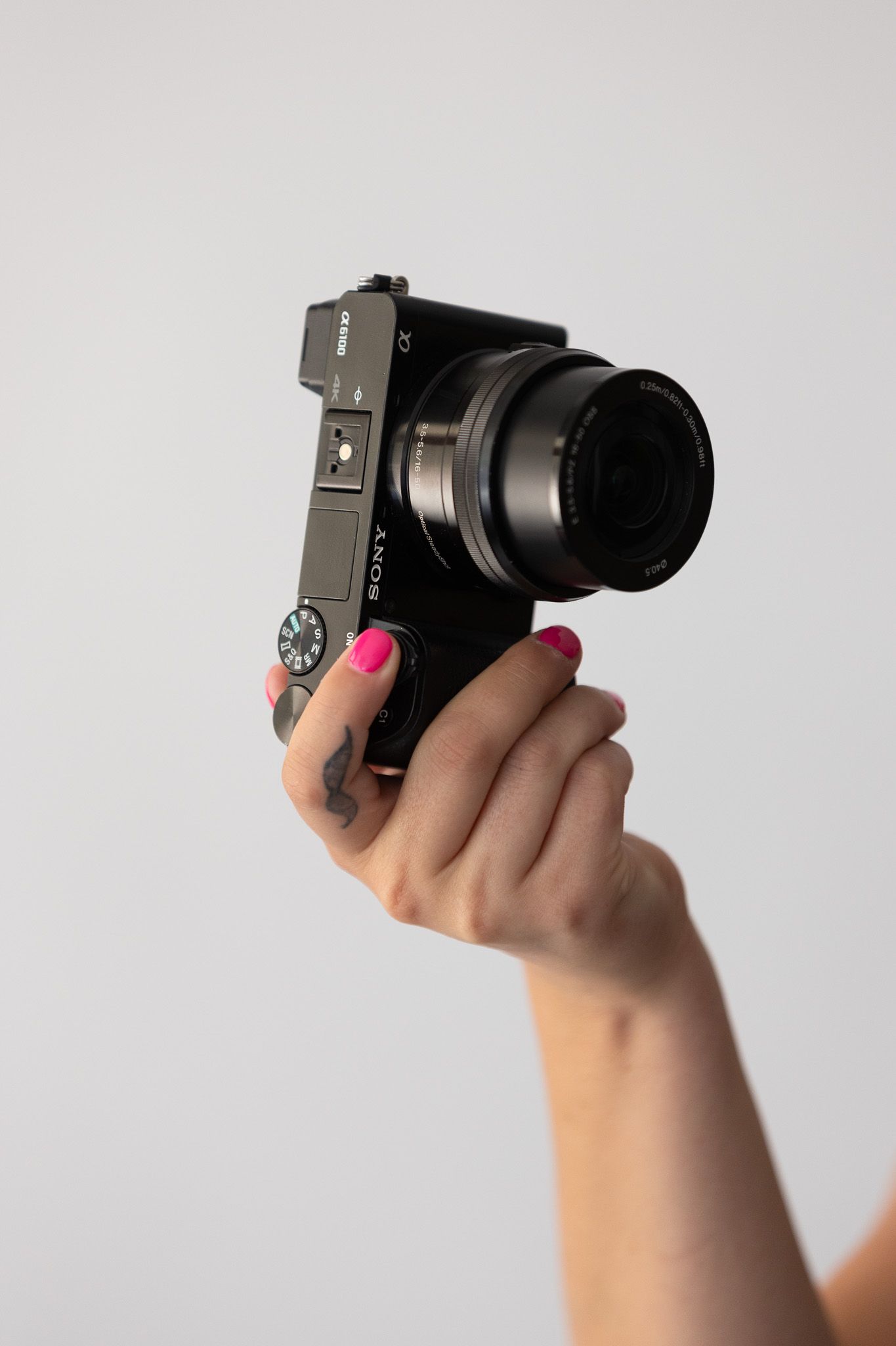 Female hand with pink nail polish holding Sony mirrorless camera with lens against clean white background.