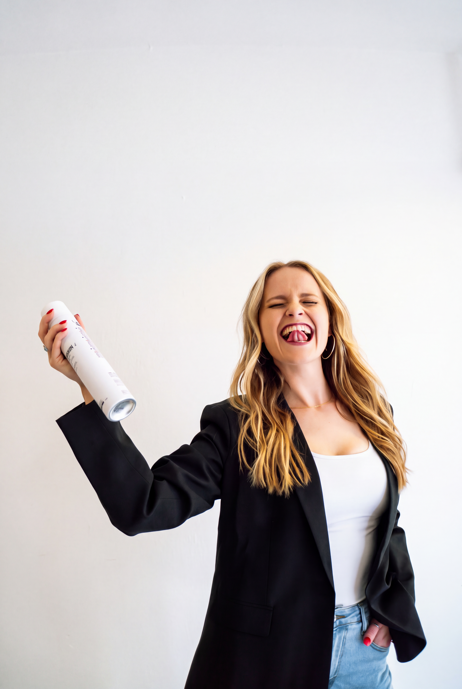 Laughing woman with long wavy blonde hair in a black blazer and white top holds a can of hair spray against a plain white background.