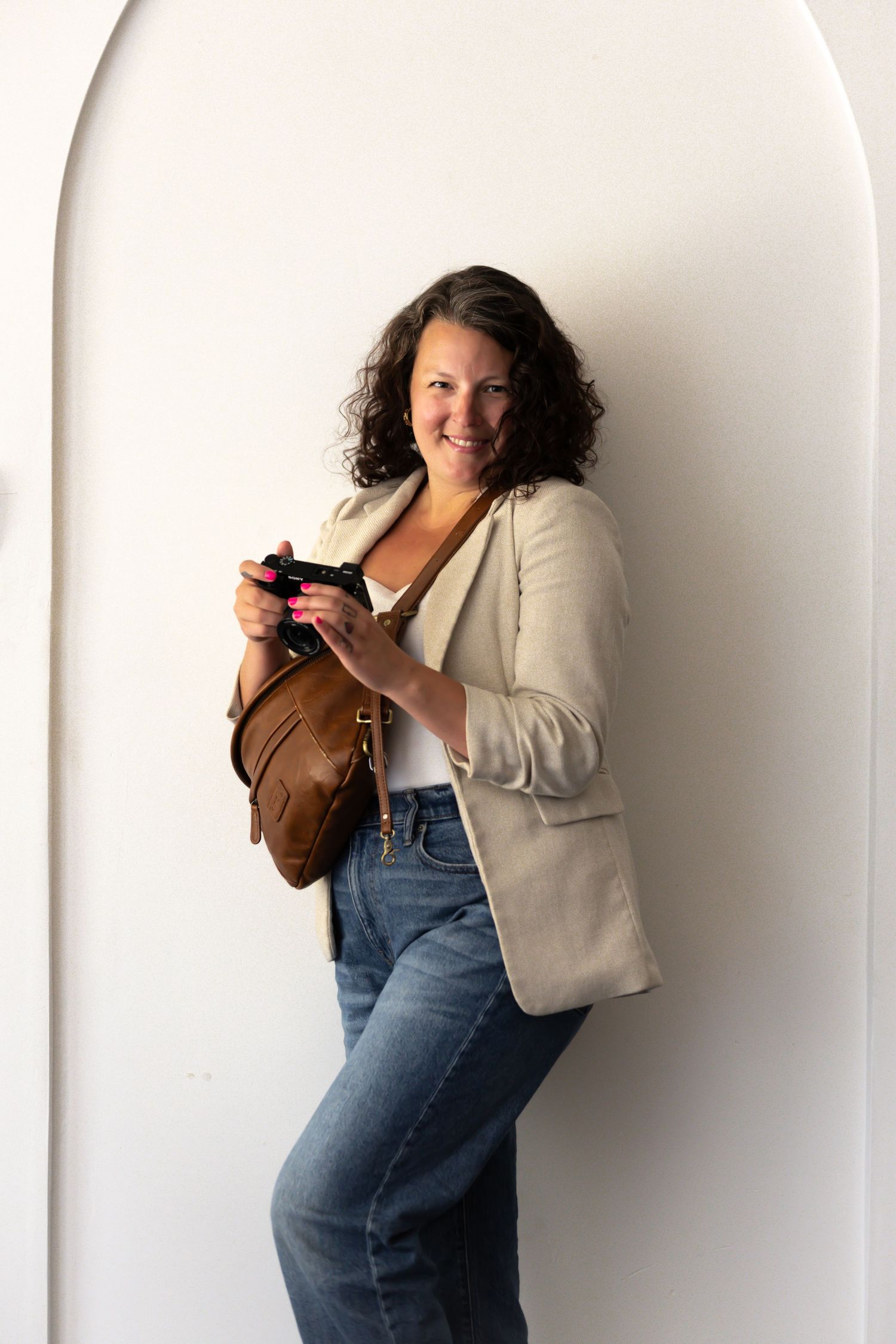 Smiling woman in beige blazer and jeans holding a camera, leaning against a white arched wall with a brown leather bag.