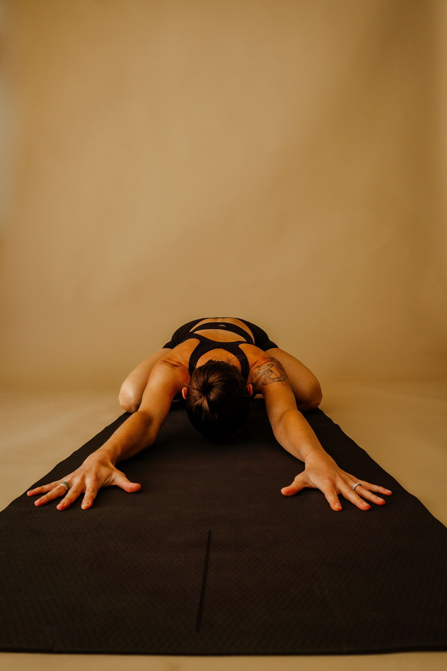 Woman in black activewear performing a deep forward fold yoga pose on a black mat against a beige background.