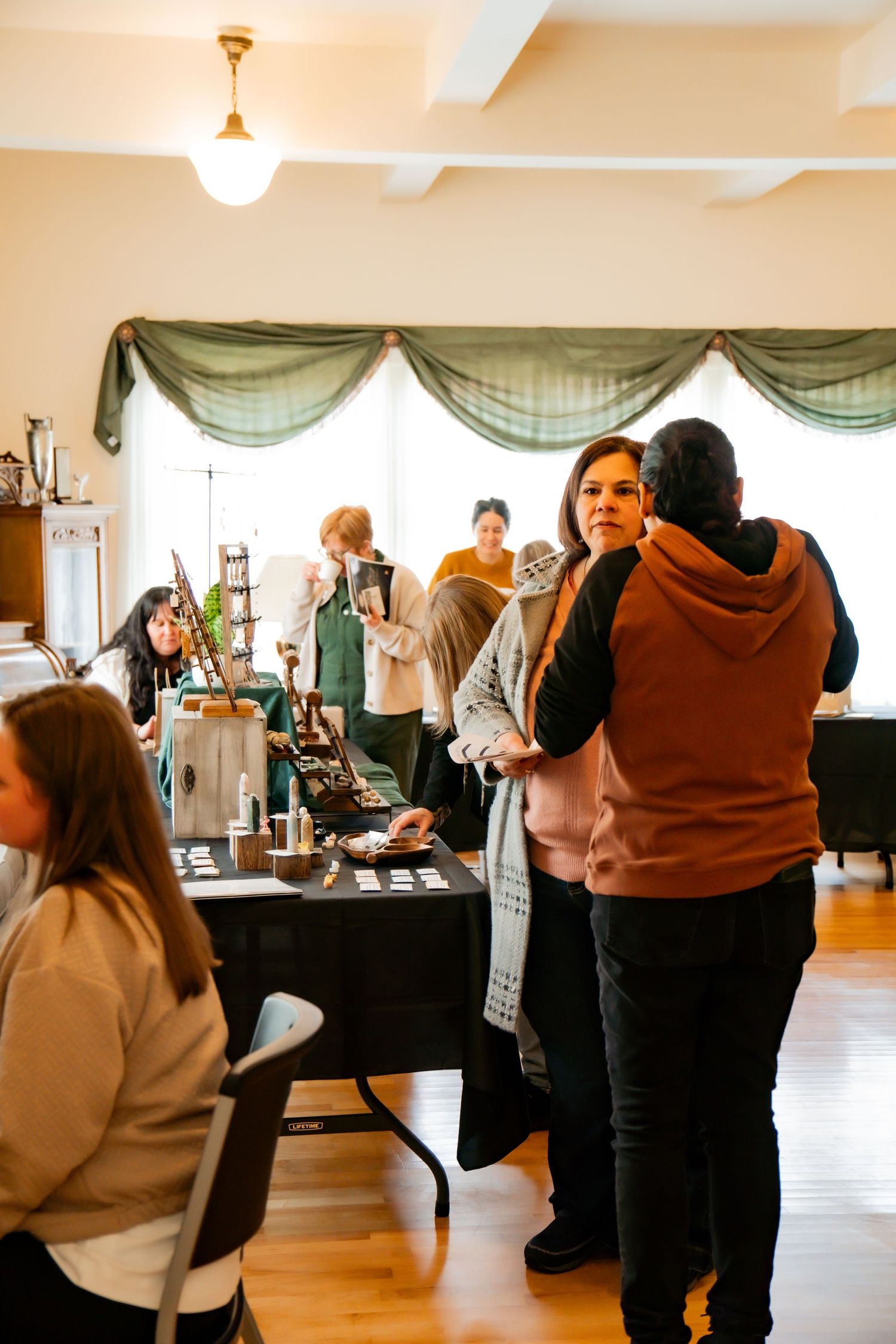 Shoppers browse handmade goods at vendor tables during a bright indoor craft market with hardwood floors and green drapes.