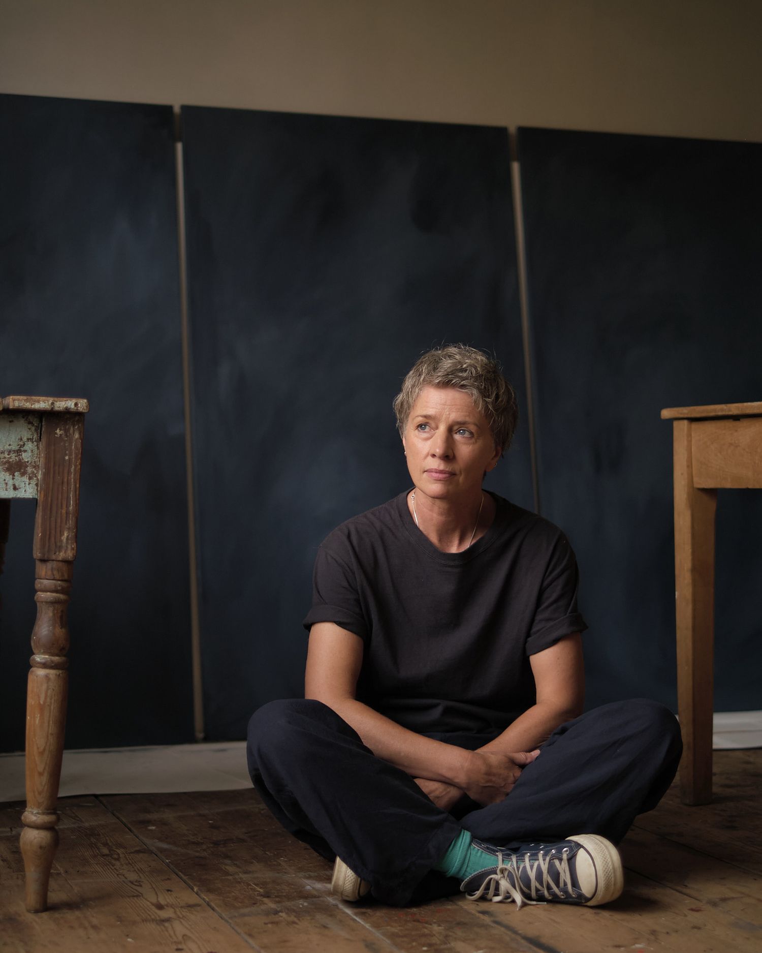 Artist sitting cross-legged on wooden floor in front of dark triptych panels in a studio setting.