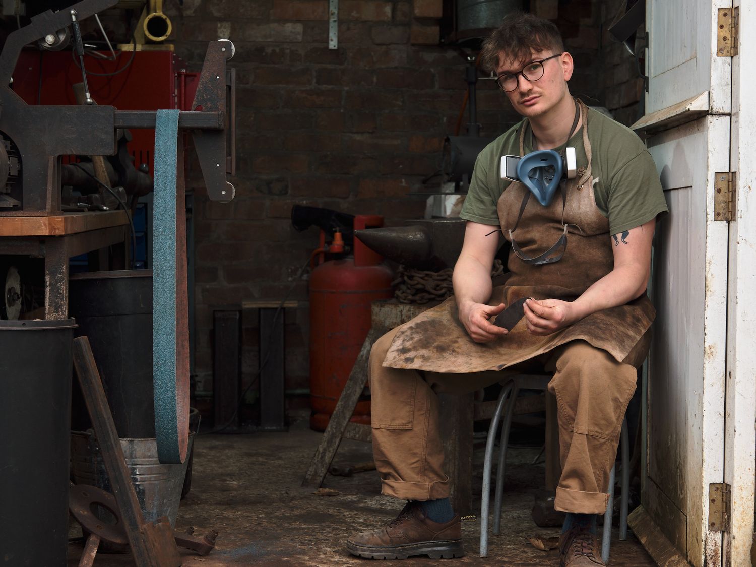 Young craftsman in apron and glasses works with leather in a rustic workshop surrounded by tools and equipment.