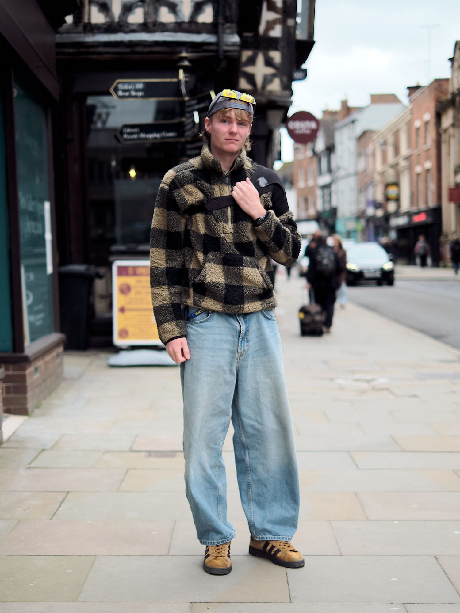 Young man in plaid fleece jacket, wide-leg jeans, and hiking boots posing on a UK high street sidewalk.