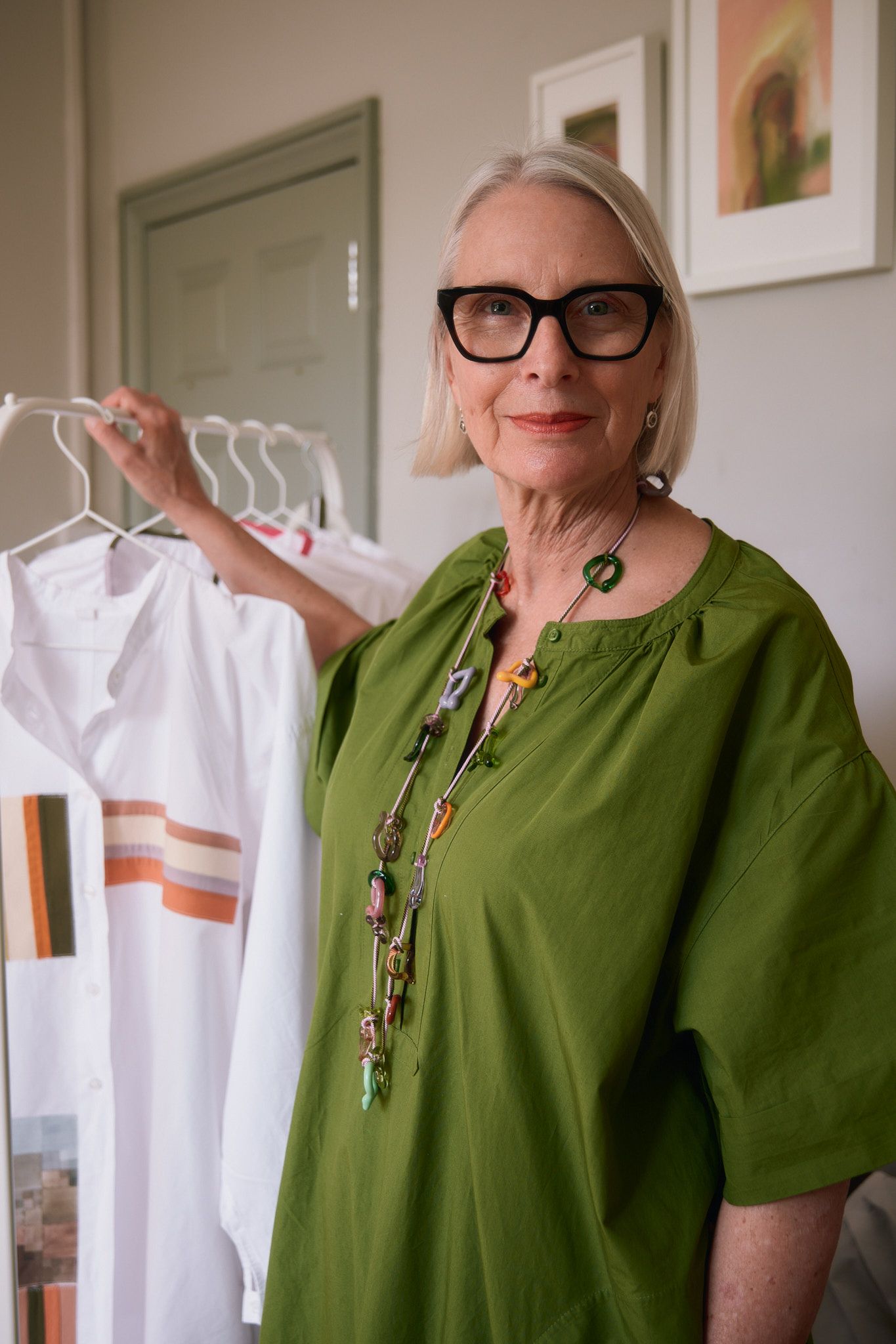 Stylish senior woman in green dress and bold jewelry holding white garments on hangers indoors.