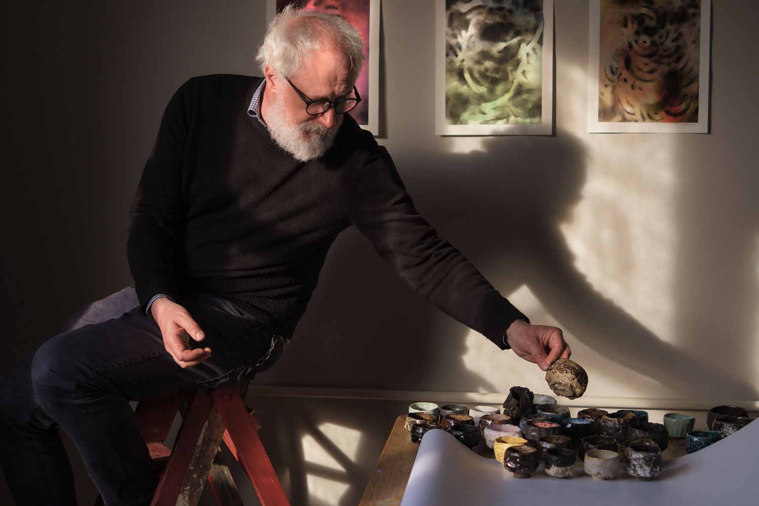 Elderly man with white beard and glasses reaching for paint bottles on a table in a sunlit art studio.