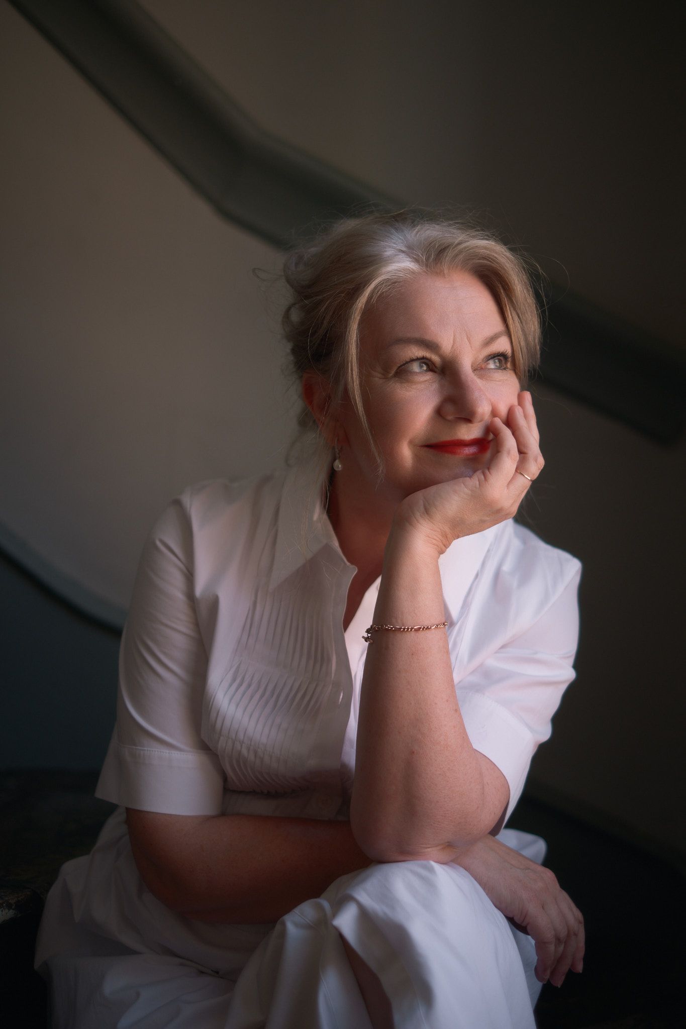Elegant older woman in white outfit resting chin on hand, gazing thoughtfully in soft light.