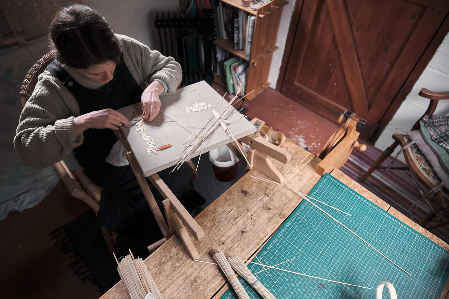 A woman weaves on a traditional wooden loom in a cozy workshop filled with books and craft supplies.