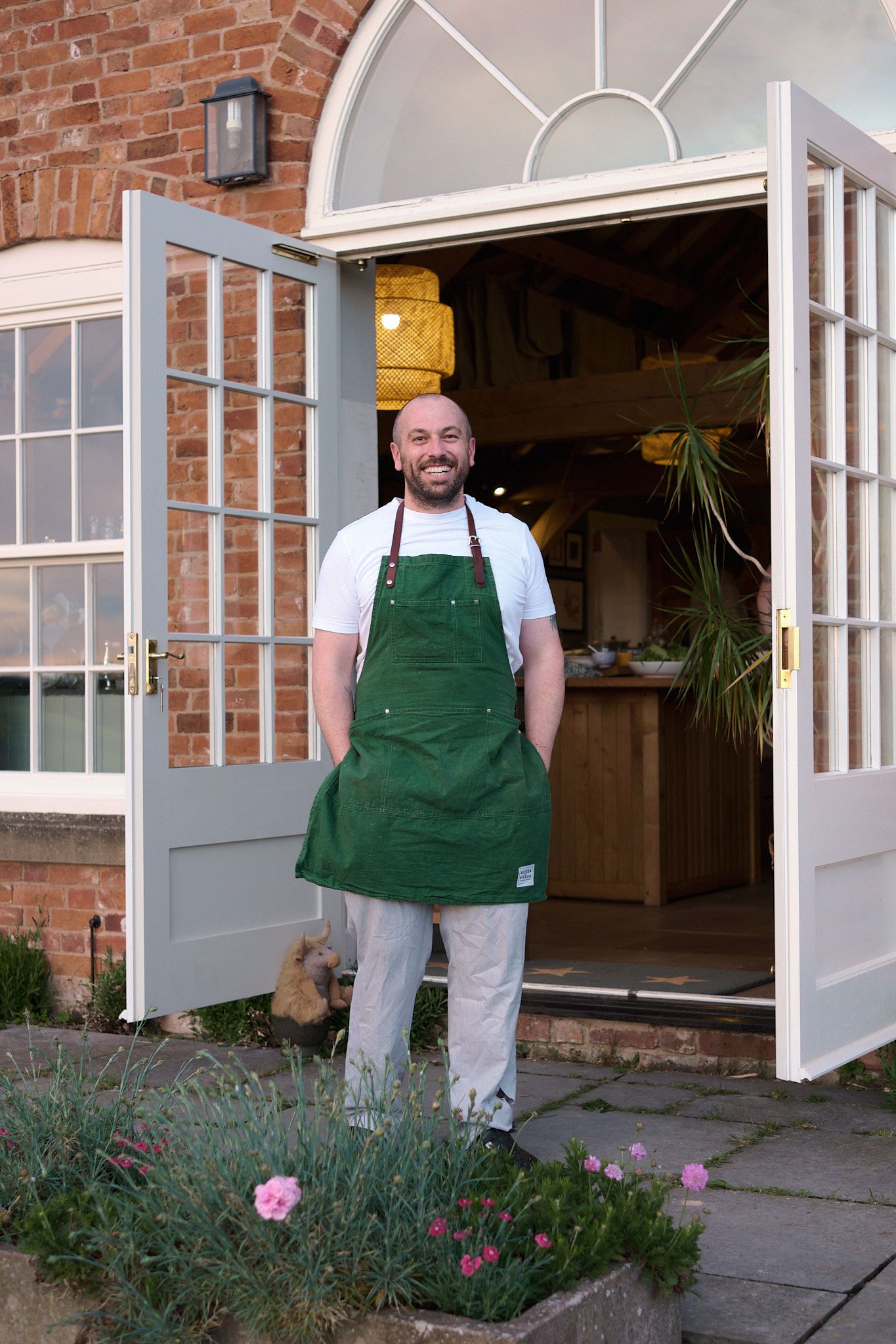 Smiling bald man in green apron stands at open French doors of a brick building.