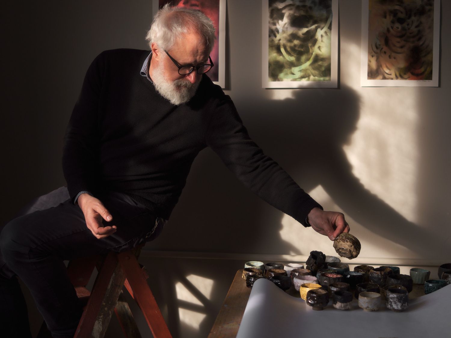Elderly bearded artist examines artwork on a table in a sunlit studio with paintings displayed on the wall.