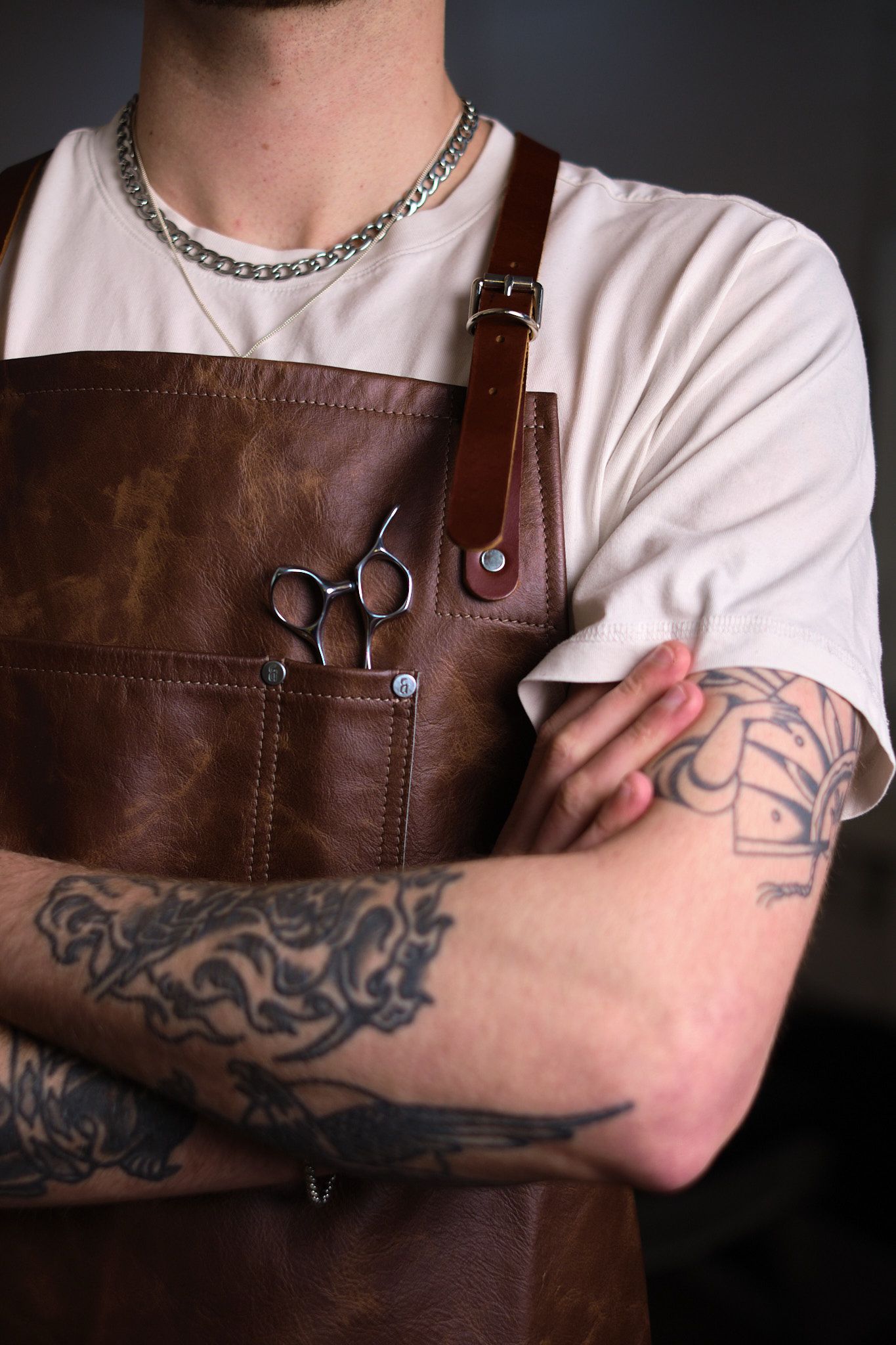 Tattooed barber in brown leather apron with scissors, arms crossed, wearing chain necklace.