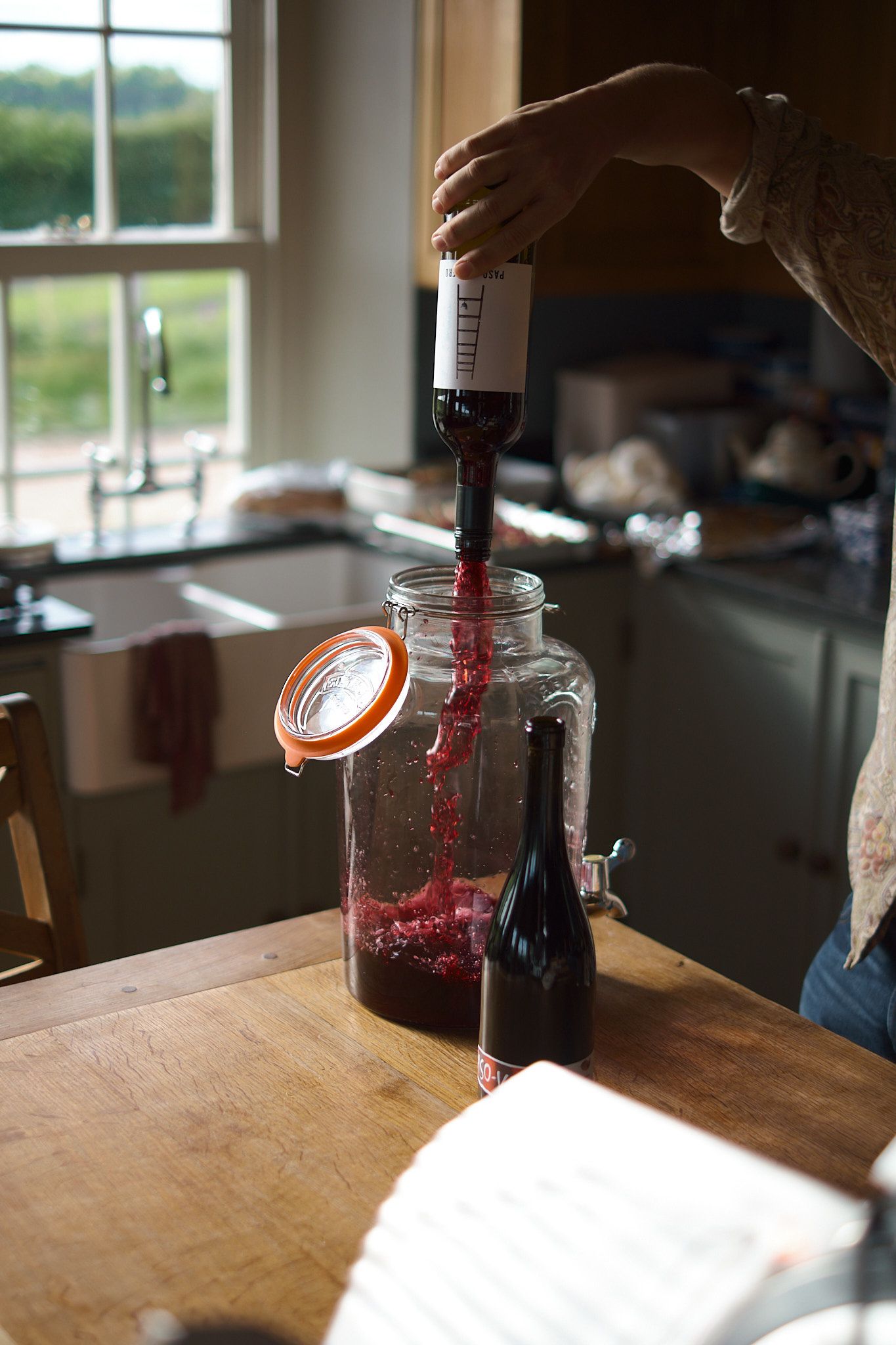 Person pouring red wine into a large glass jar on a wooden kitchen counter.