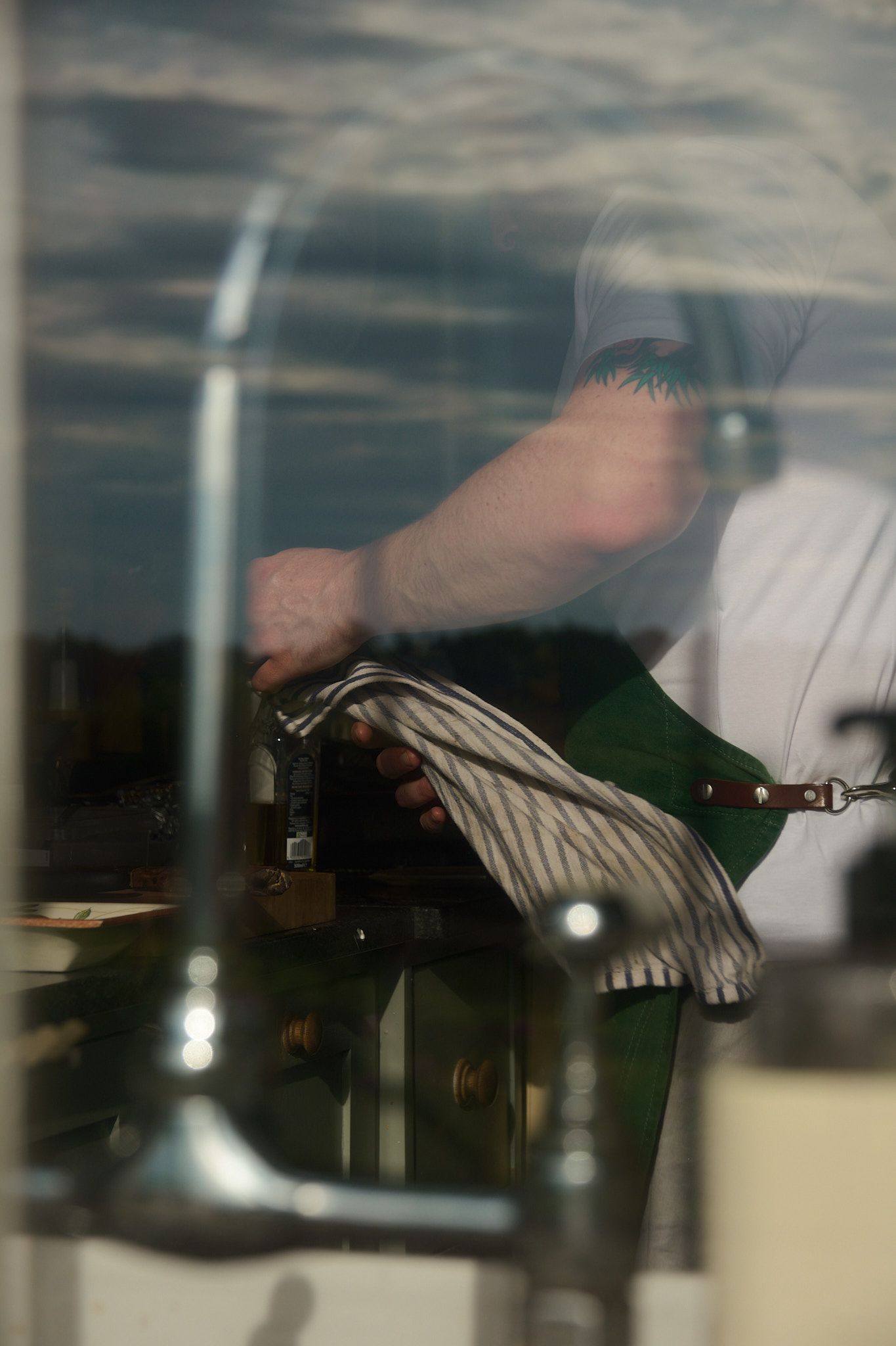 A chef in a white shirt and green apron polishes a surface seen through a reflective window.
