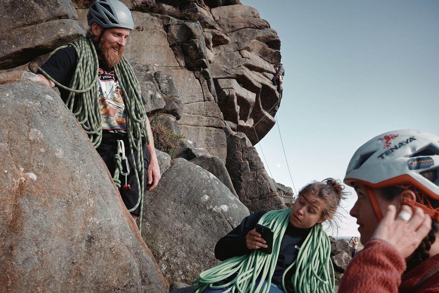 Three rock climbers with helmets and green ropes prepare for a climb on a rocky mountain face outdoors.