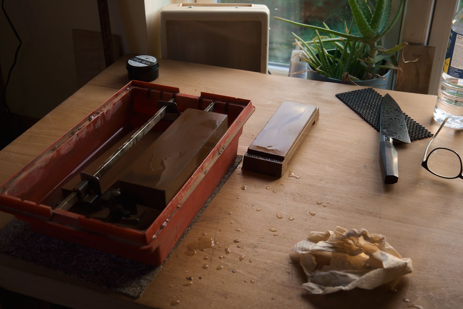 A wooden workbench with a knife sharpening kit, whetstone, chef knife, and scattered wood shavings near a window.