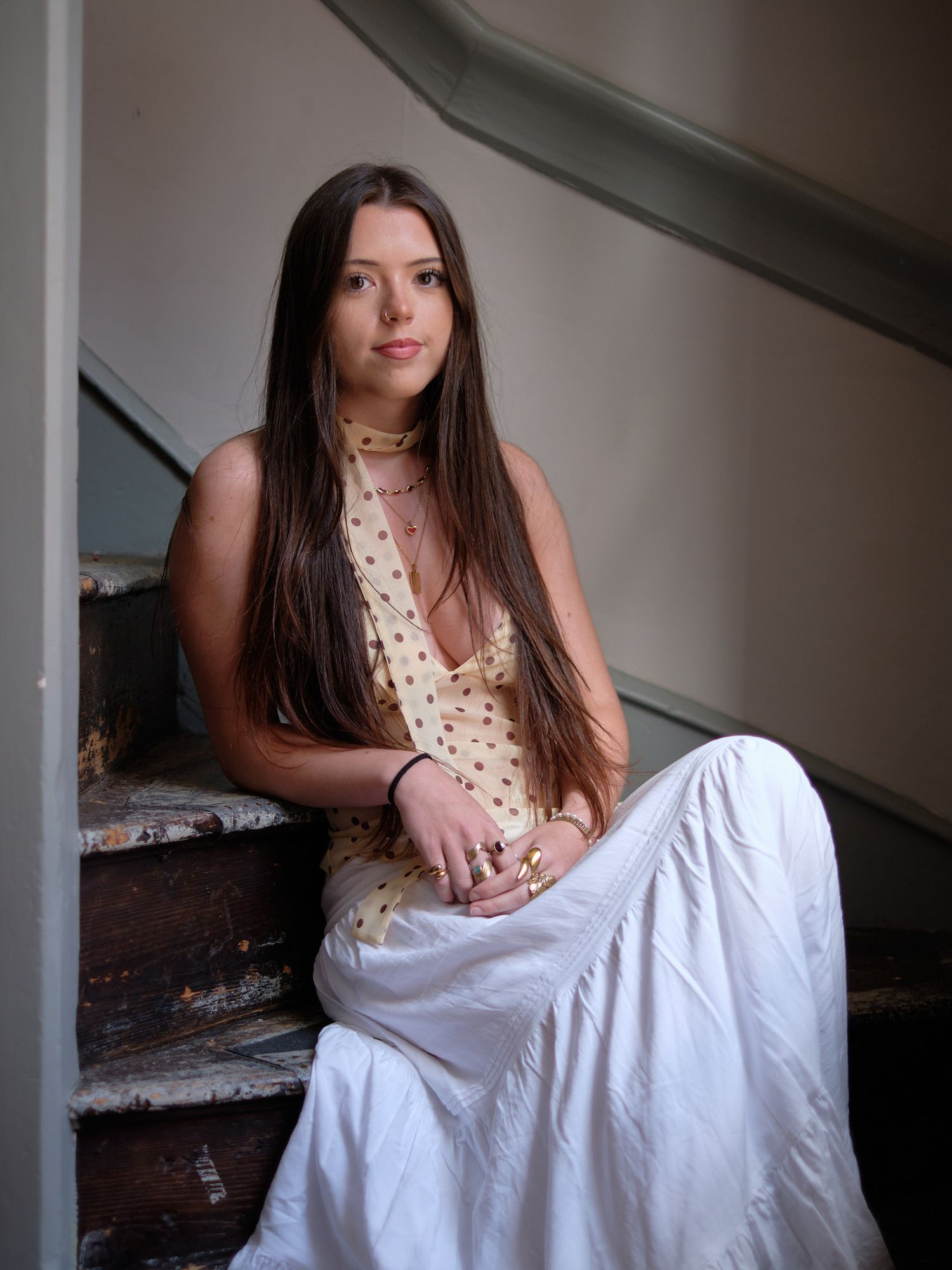 Young woman with long dark hair wearing a floral top and white skirt seated gracefully on old wooden stairs.