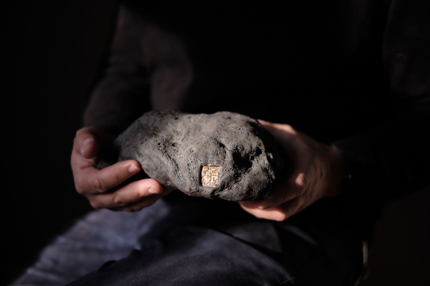 Hands holding a large grey rock with a small embedded fossil against a dark background.