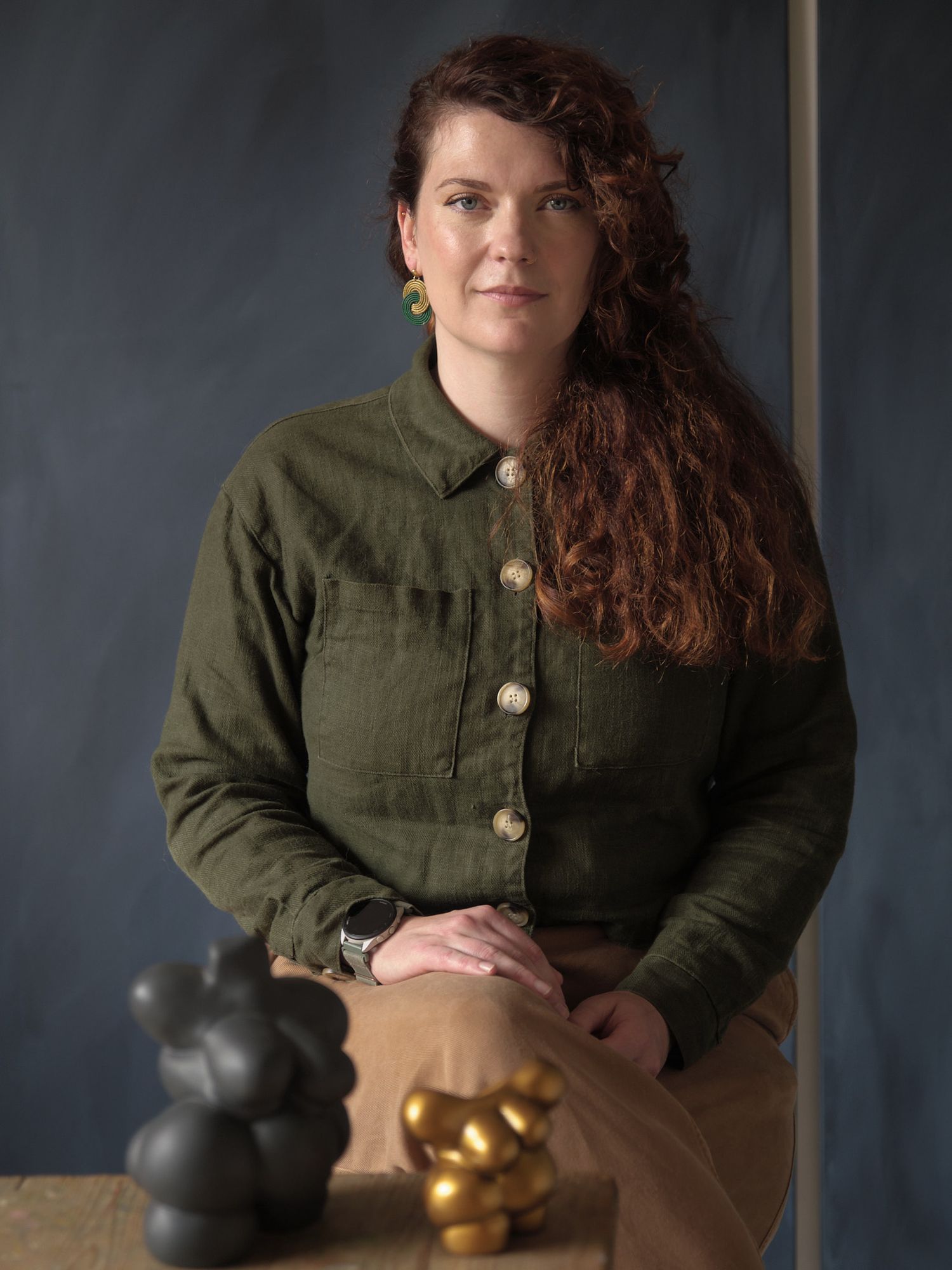 Woman in green linen shirt poses with small black and gold sculptures on a wooden table against a gray background.