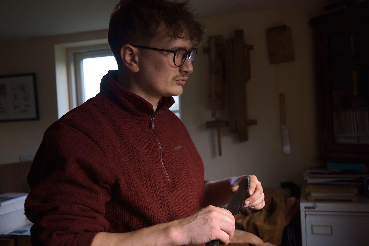 Young man in red fleece jacket working with his hands in a dimly lit workshop.