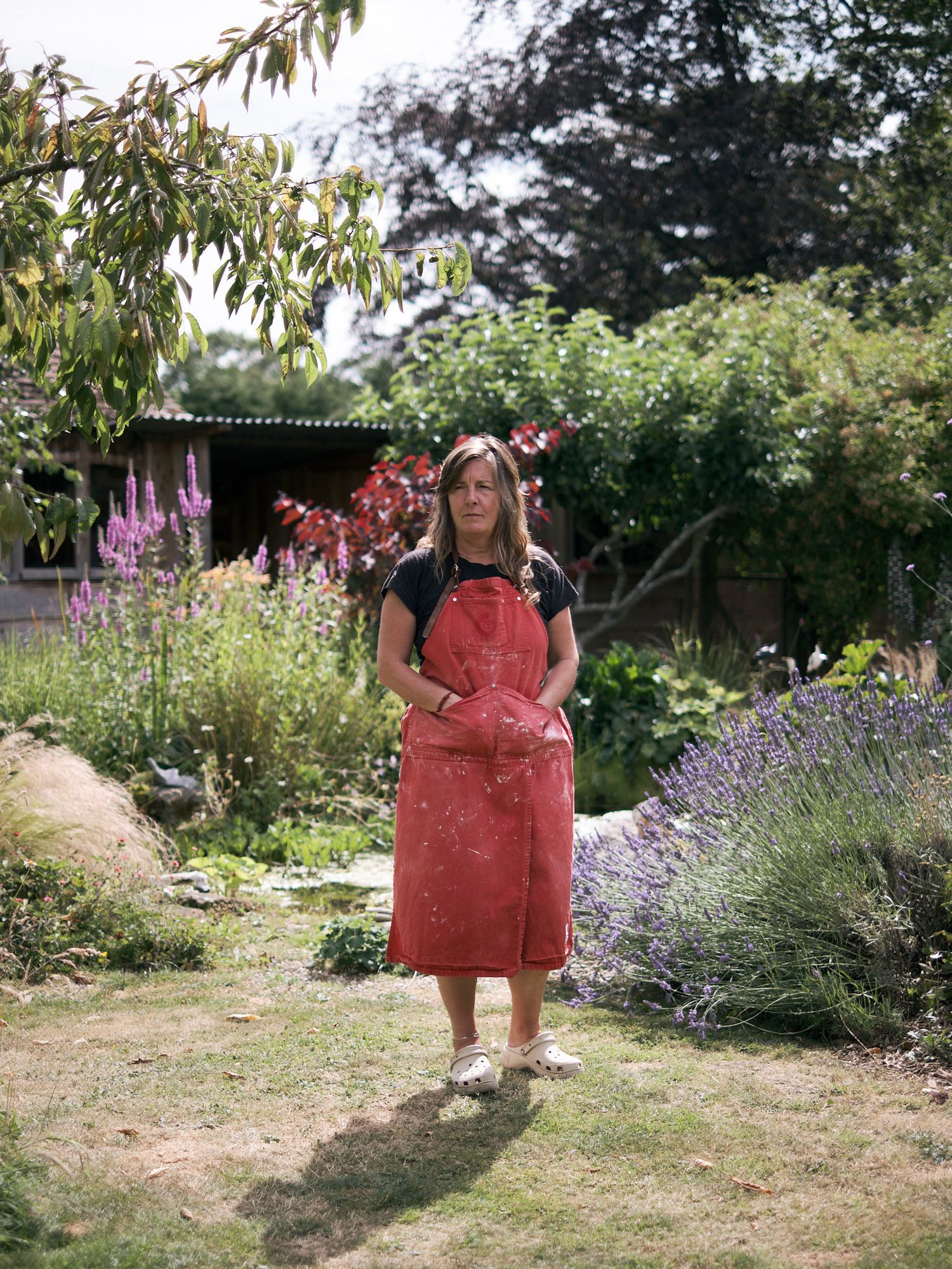Female potter in red apron standing in a blooming garden with lavender and roses on sunny day.