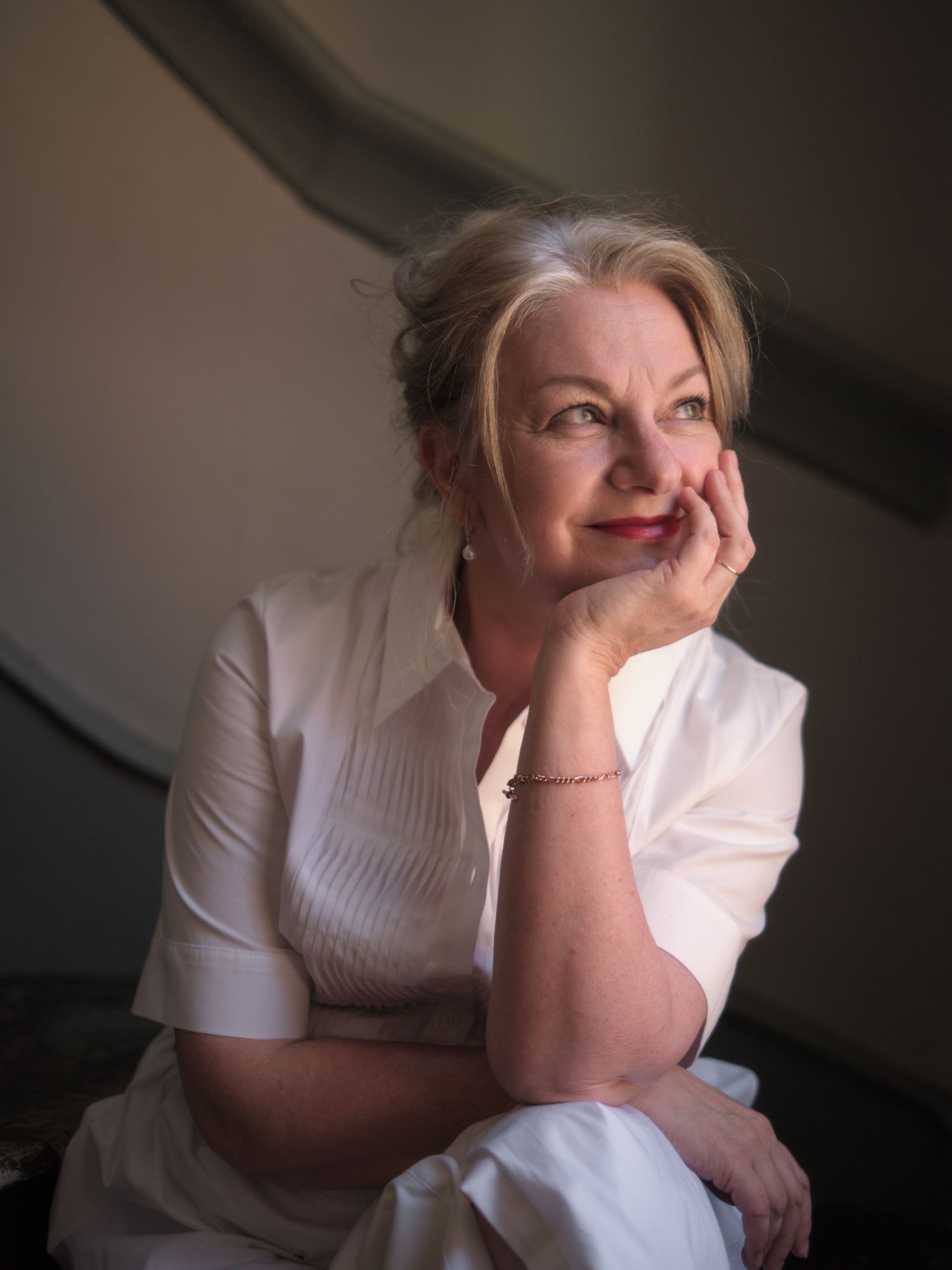 A thoughtful middle-aged woman in white clothing rests her chin on her hand, gazing upward in a softly lit portrait.
