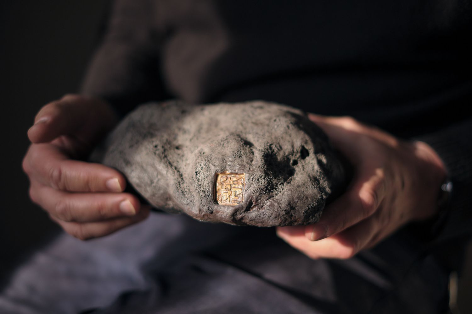 Hands holding a large gray rock with a small golden seal embedded in its surface.