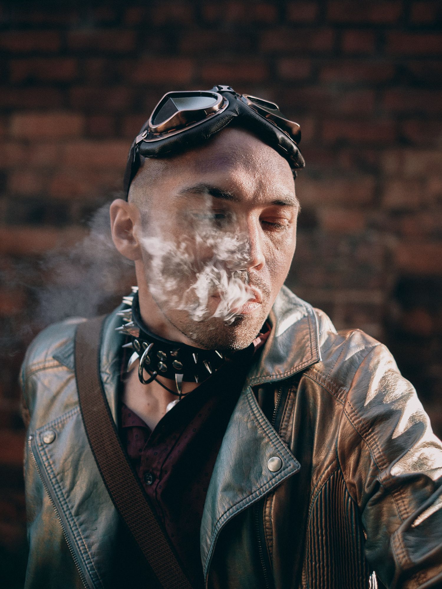 Edgy punk man wearing goggles, spiked collar, and leather jacket blowing smoke in front of brick wall.