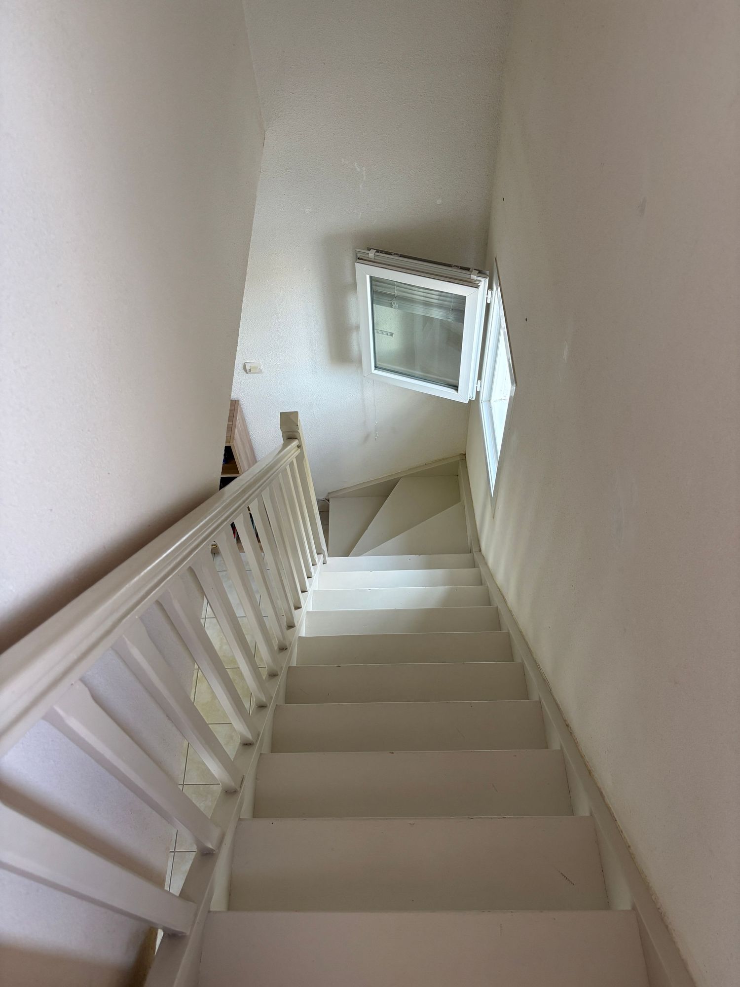 Escalier intérieur blanc à rampe en bois, éclairé par une petite fenêtre