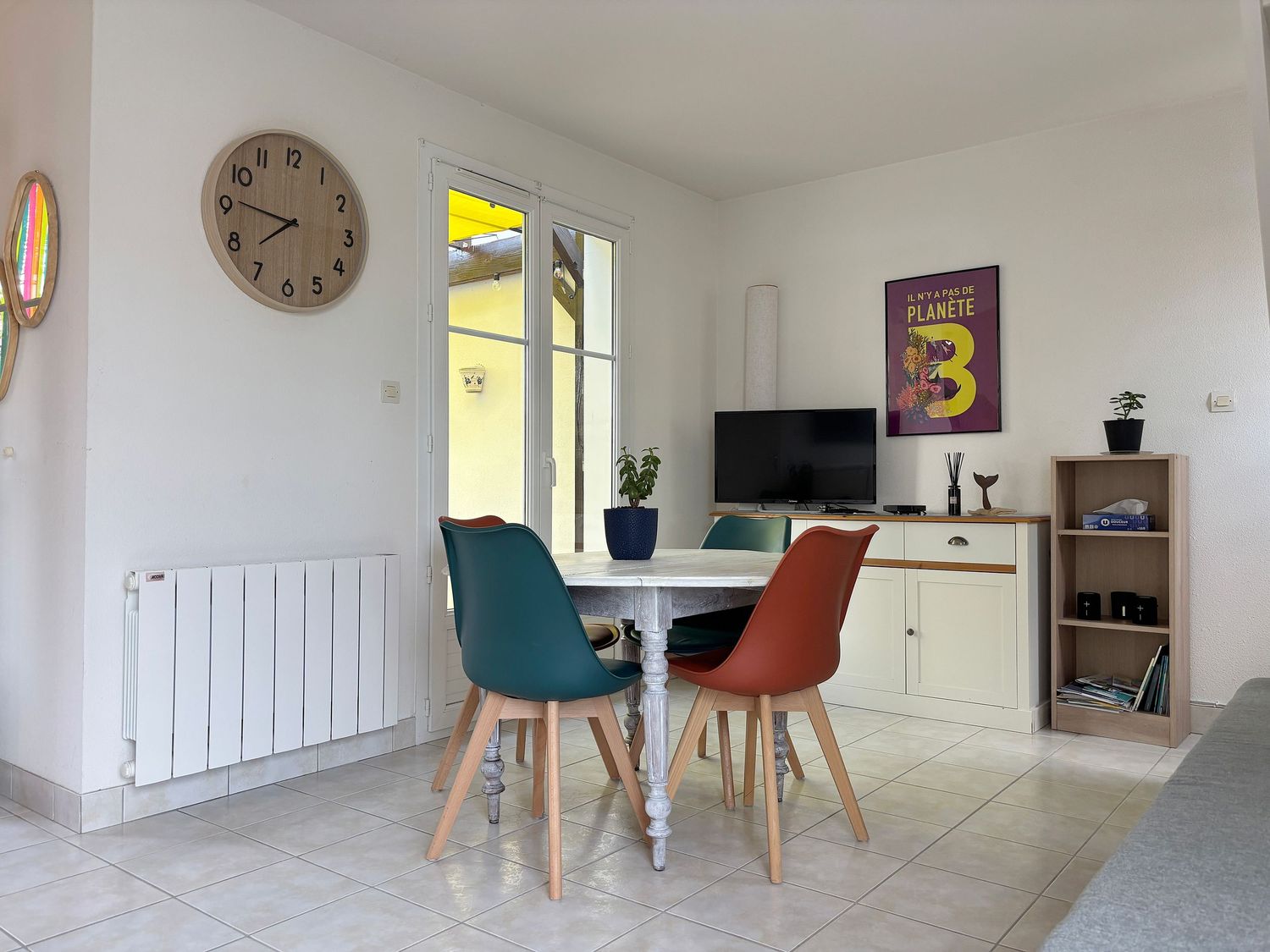 Séjour ouvert lumineux avec table ronde, télévision, chaises colorées, escalier et horloge
