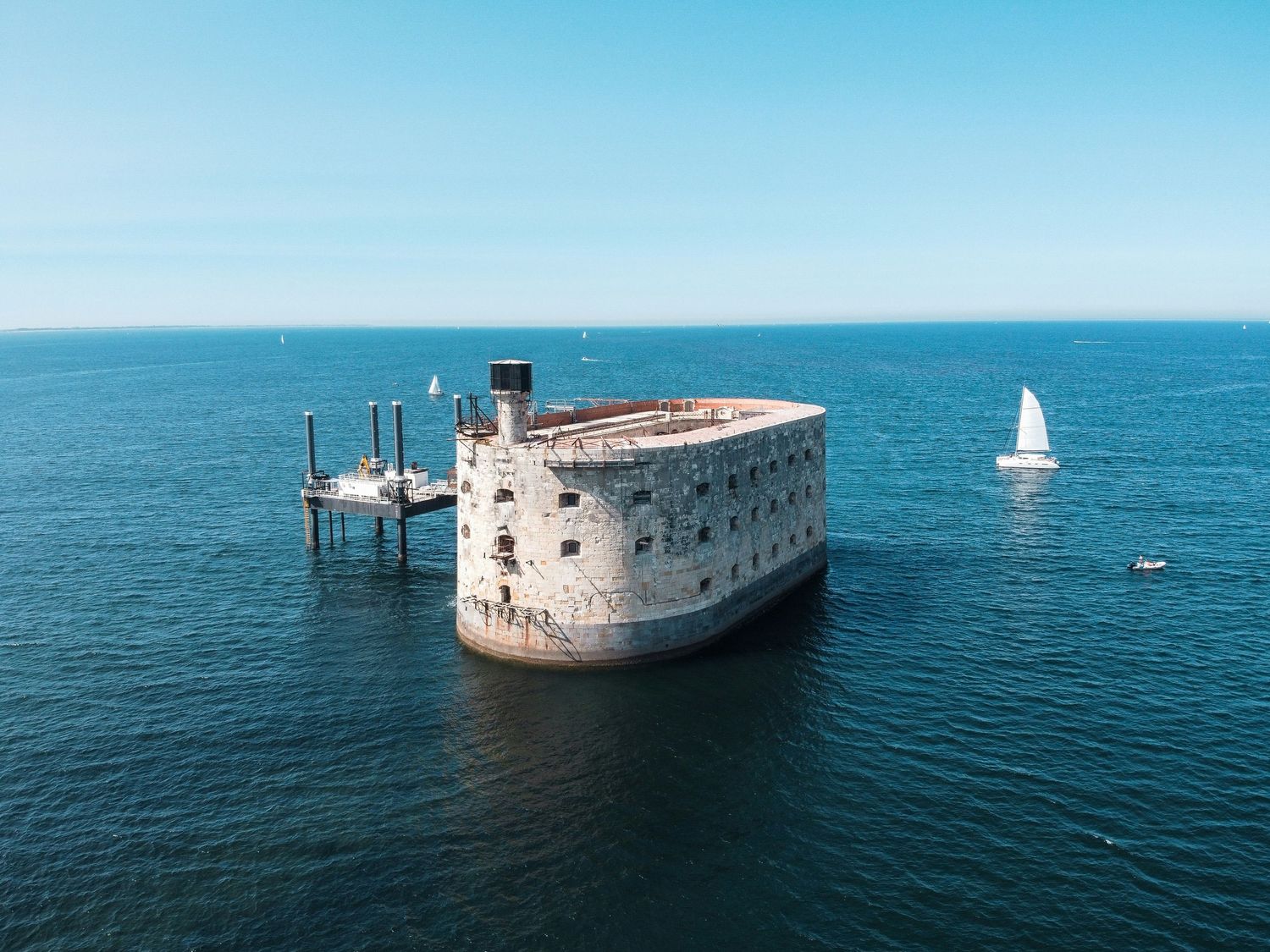 Aerial view of Fort Boyard stone fortress surrounded by blue ocean waters with a sailboat nearby.