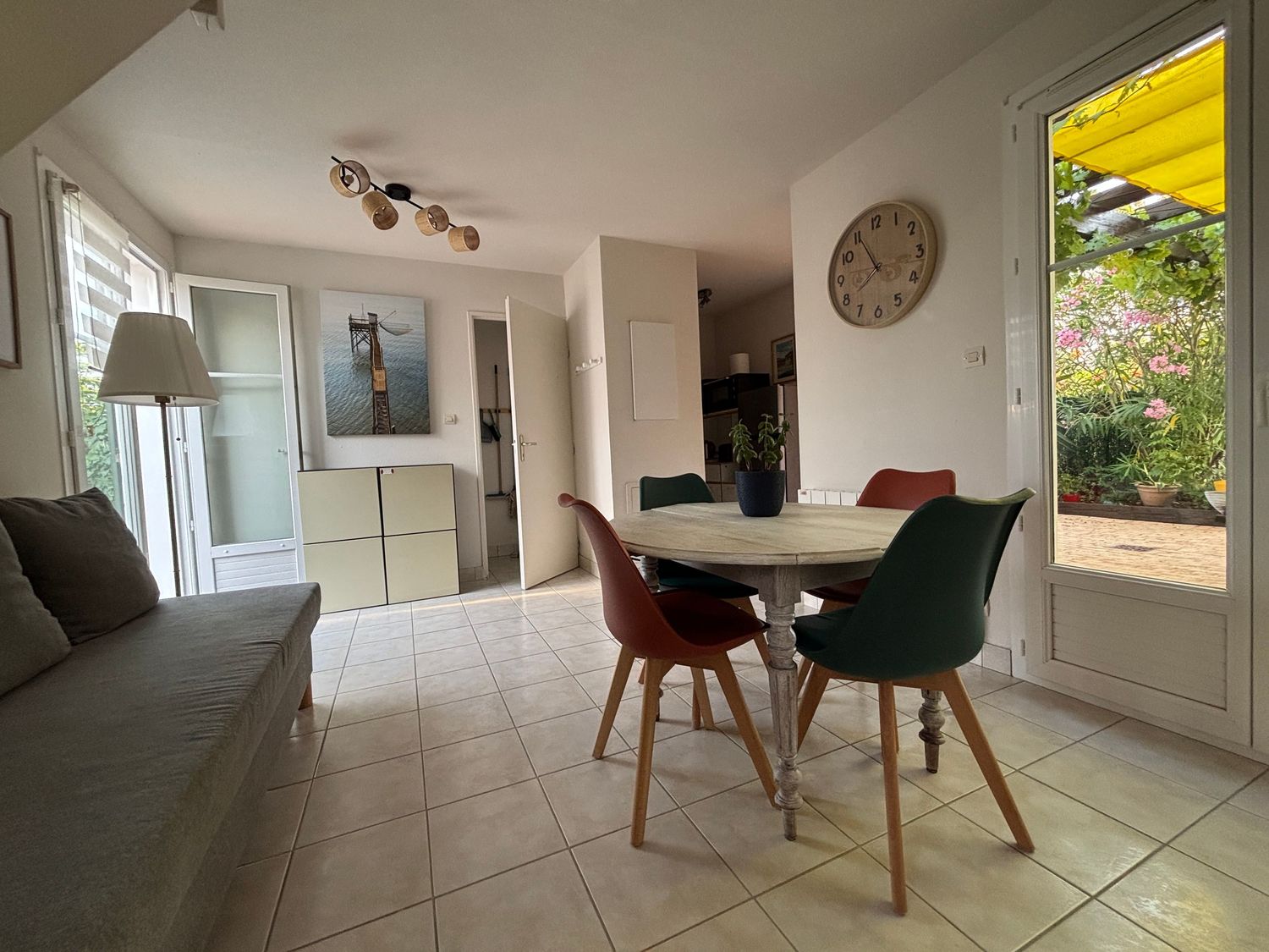 Coin repas lumineux avec chaises colorées, table ronde, escalier et horloge murale, vue sur la terrasse extérieure de la maison de Fouras