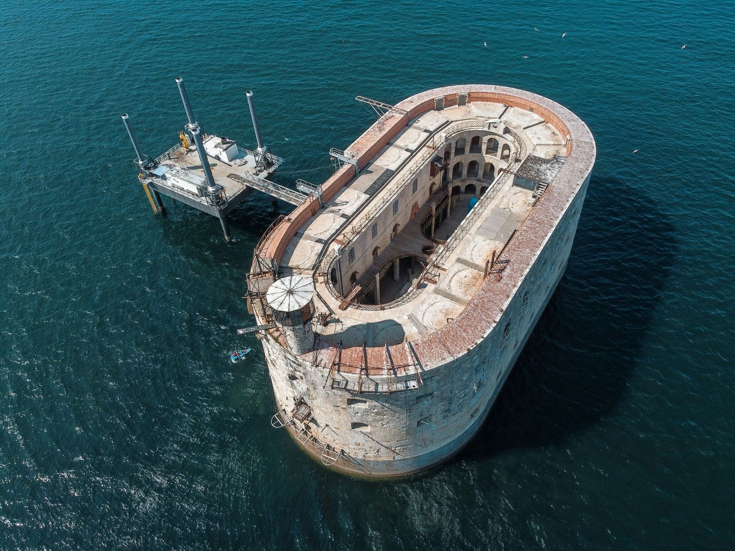 Aerial view of an abandoned oval sea fort surrounded by turquoise ocean waters, featuring rusty walls and a small dock.