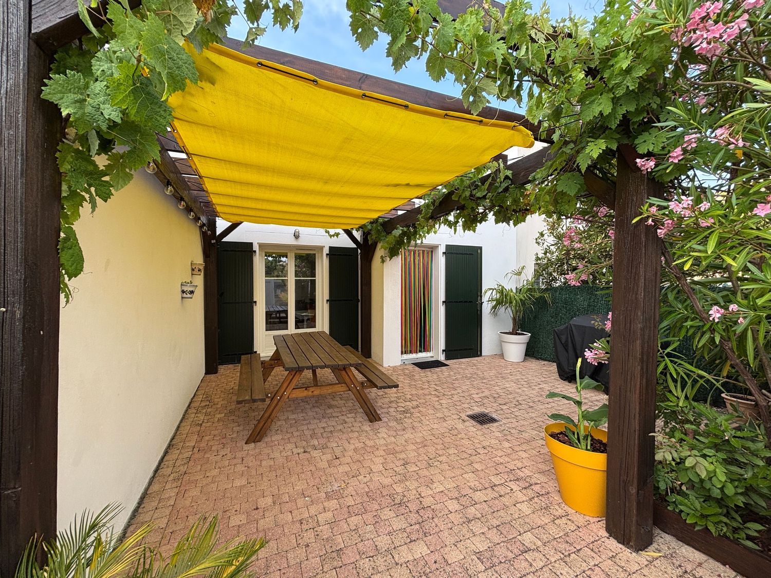 Sunny patio with yellow shade sail, wooden picnic table, and lush garden plants on brick pavers.