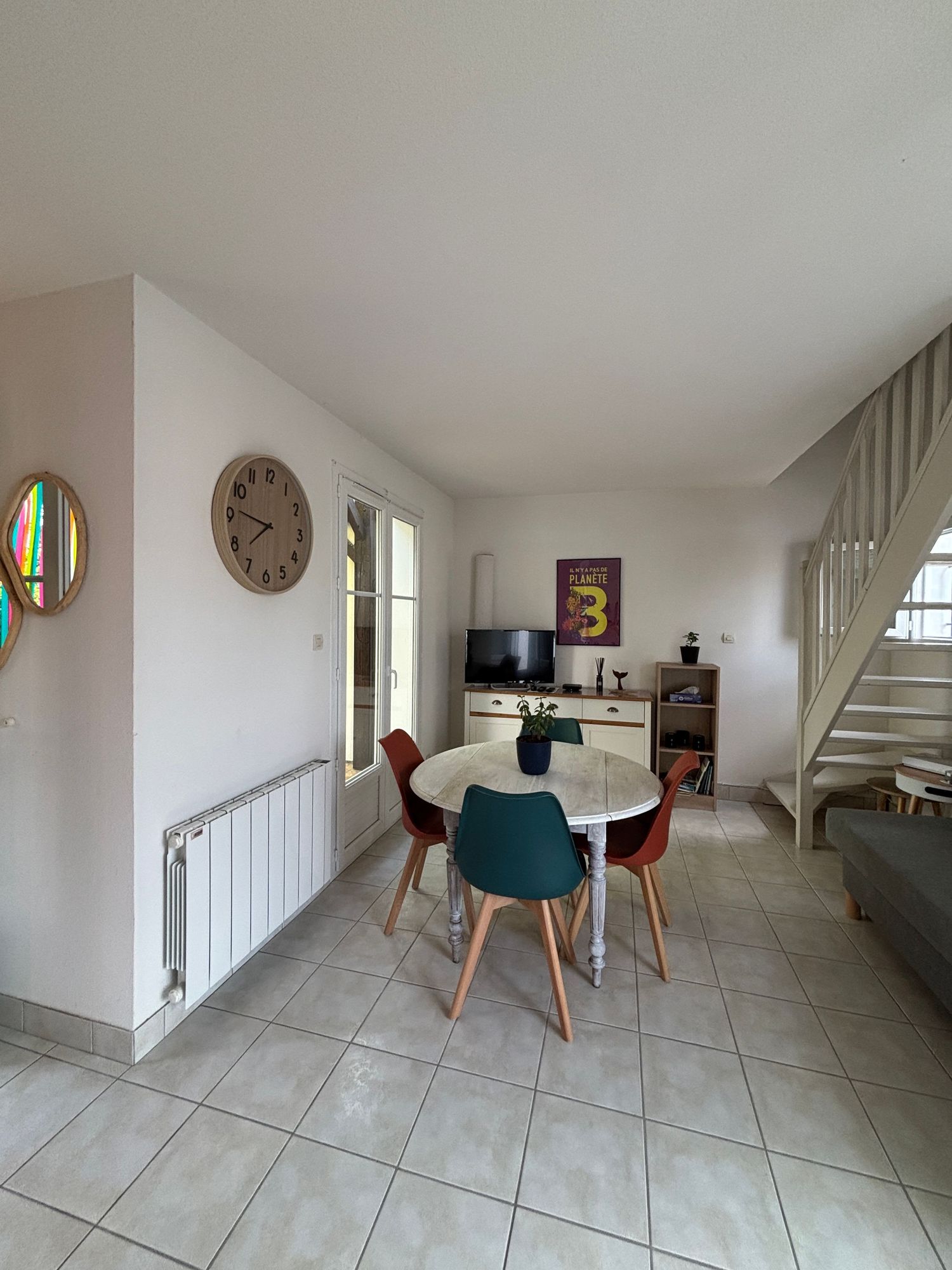 Coin repas lumineux avec chaises colorées, table ronde, télévision, escalier et horloge murale, point de vue intérieur