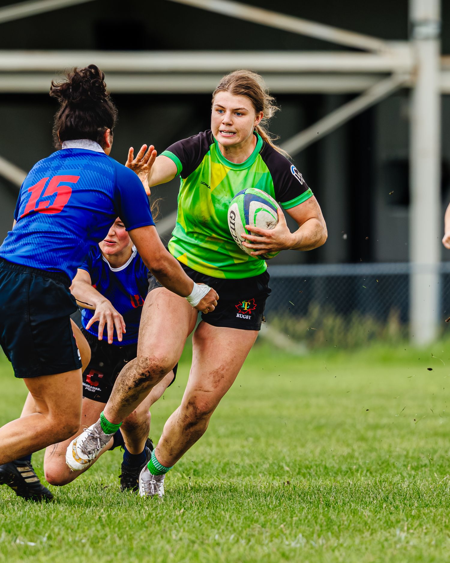 Female rugby player in green jersey charges toward defender during outdoor women's rugby match.