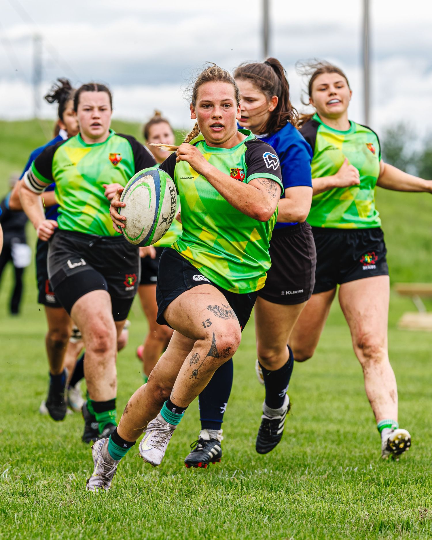 Female rugby player in green jersey runs with ball during competitive women's rugby match on grass.