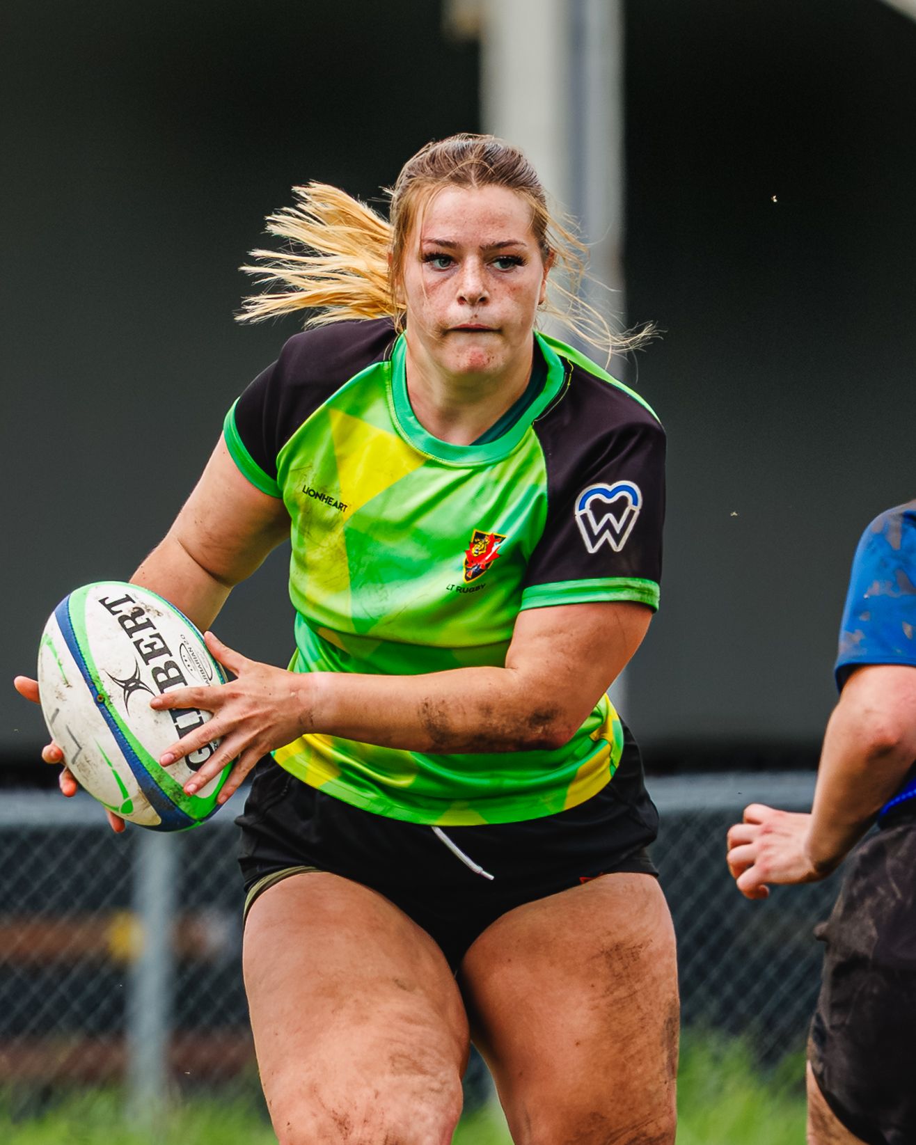 Blonde female rugby player runs with Gilbert ball in green jersey during women's rugby game.