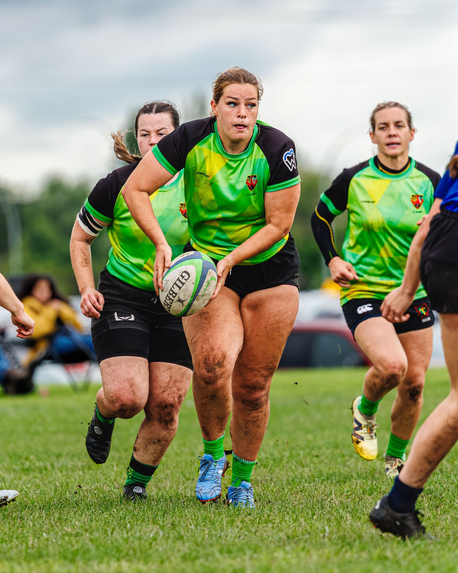 Female rugby player in green jersey carries ball while teammates support during outdoor match.