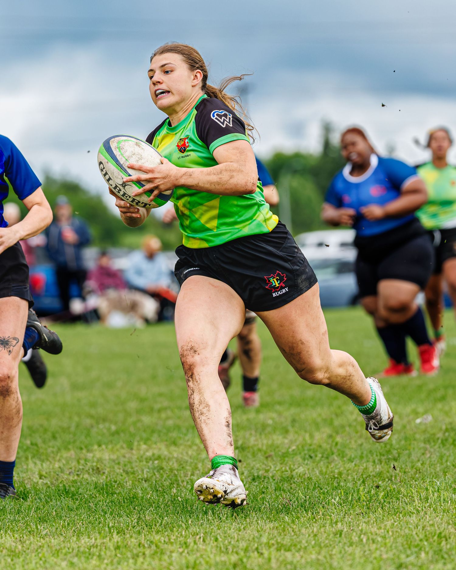 Female rugby player in green jersey sprints with ball during competitive women's rugby match.