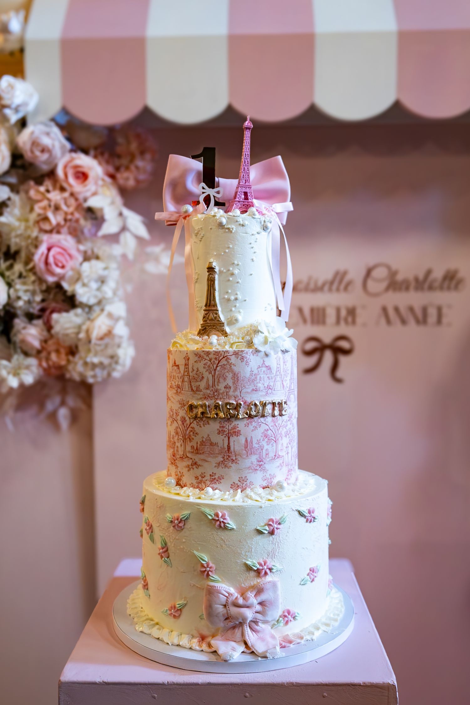 Elegant three-tier Paris-themed first birthday cake with Eiffel Tower, pink bows, floral details, and number 1 topper.