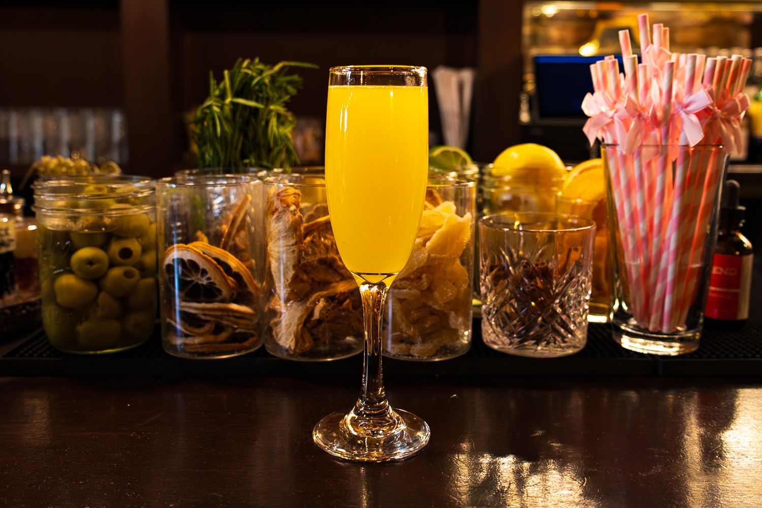 A golden mimosa in a champagne flute surrounded by bar garnishes, olives, citrus, and pink striped straws.