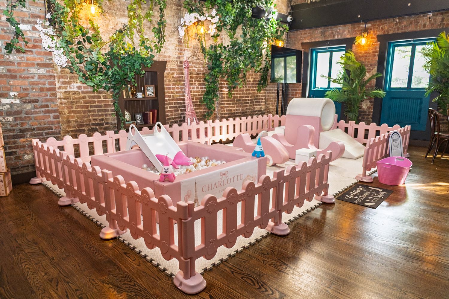 Pink picket fence ball pit play area with pastel toys inside a rustic brick-walled event space with string lights.