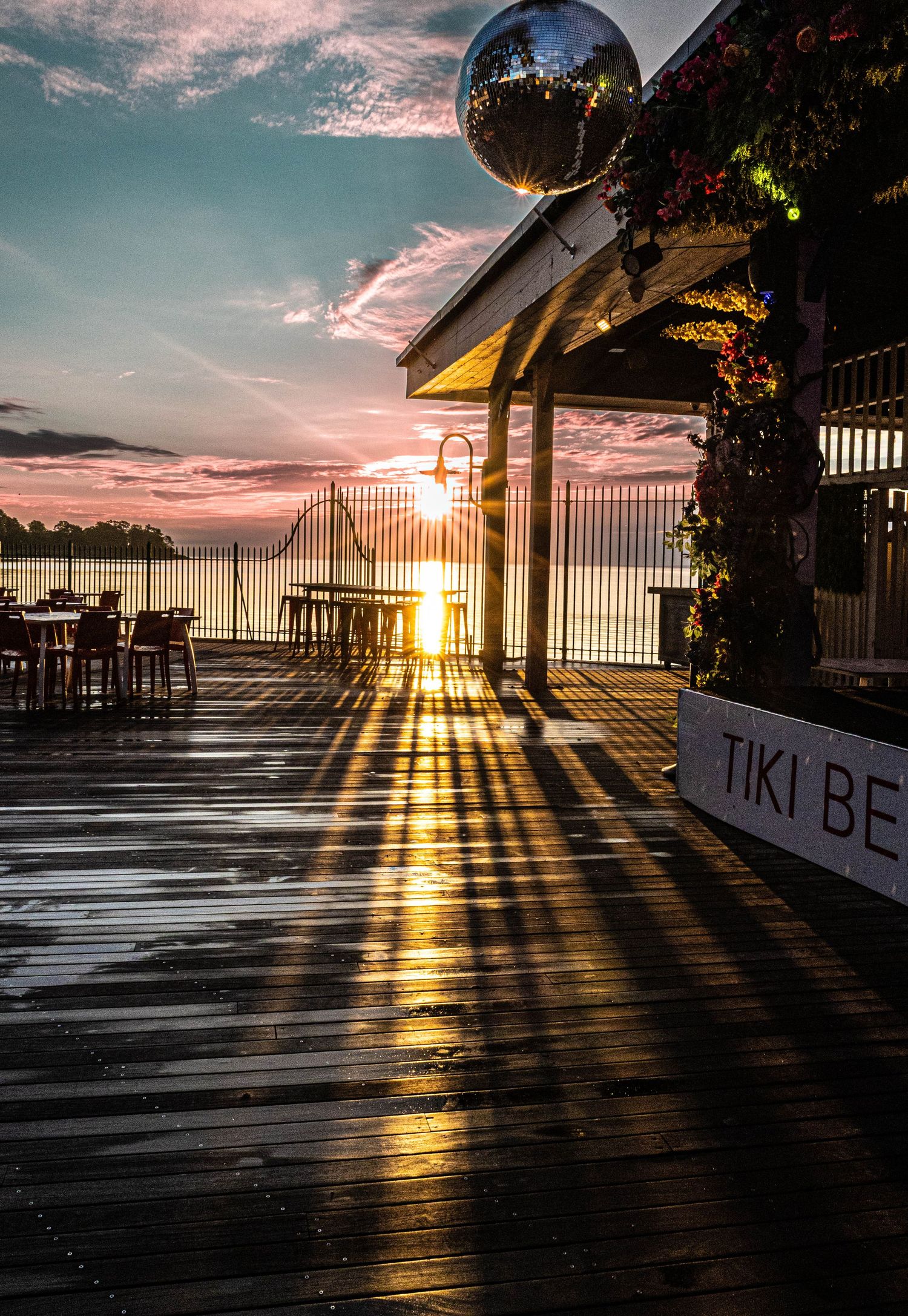 Sunset over Tiki Beach bar deck with dramatic light rays reflecting on wet wooden boards and disco ball overhead.