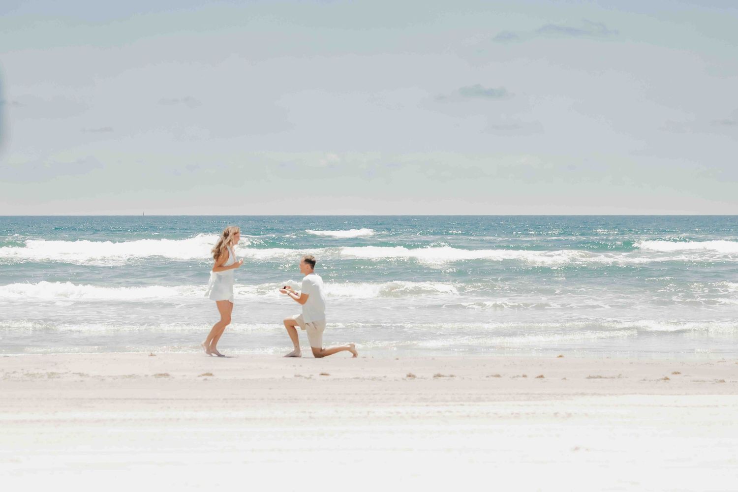 Man proposing on bended knee to woman on a white sandy beach with ocean waves in the background.