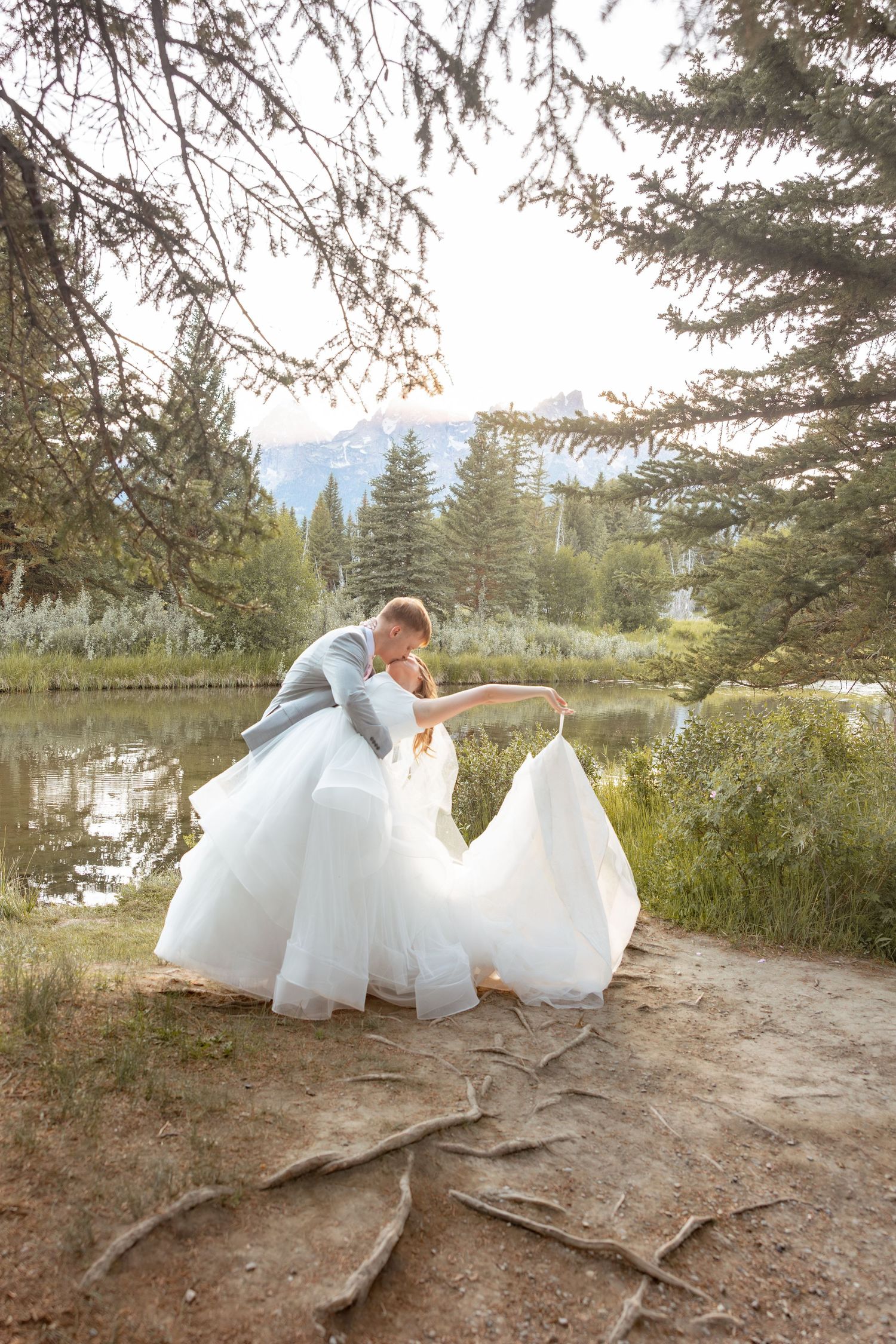 Bride and groom share a romantic dip kiss by a mountain lake surrounded by evergreen trees.