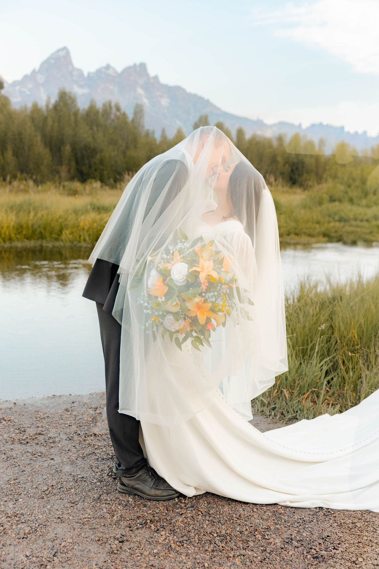 Bride and groom kissing under a veil with a colorful bouquet, Grand Teton mountains and river in the background.